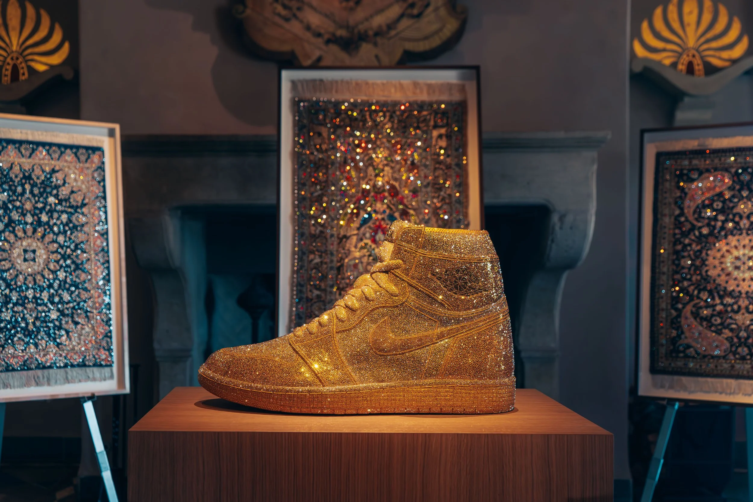 A golden, sparkly Nike high-top sneaker displayed on a wooden pedestal, surrounded by framed abstract artwork with intricate patterns in a gallery setting.