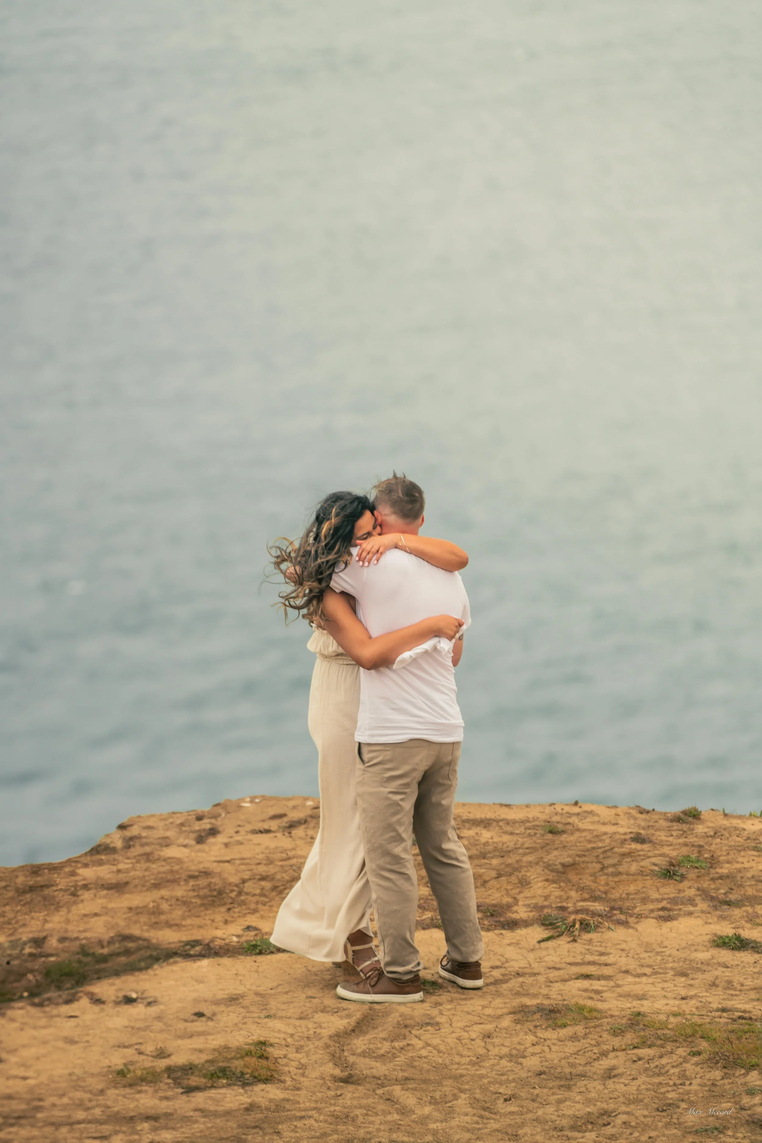 A couple embracing on a rocky cliff next to the ocean, the woman hugging the man.