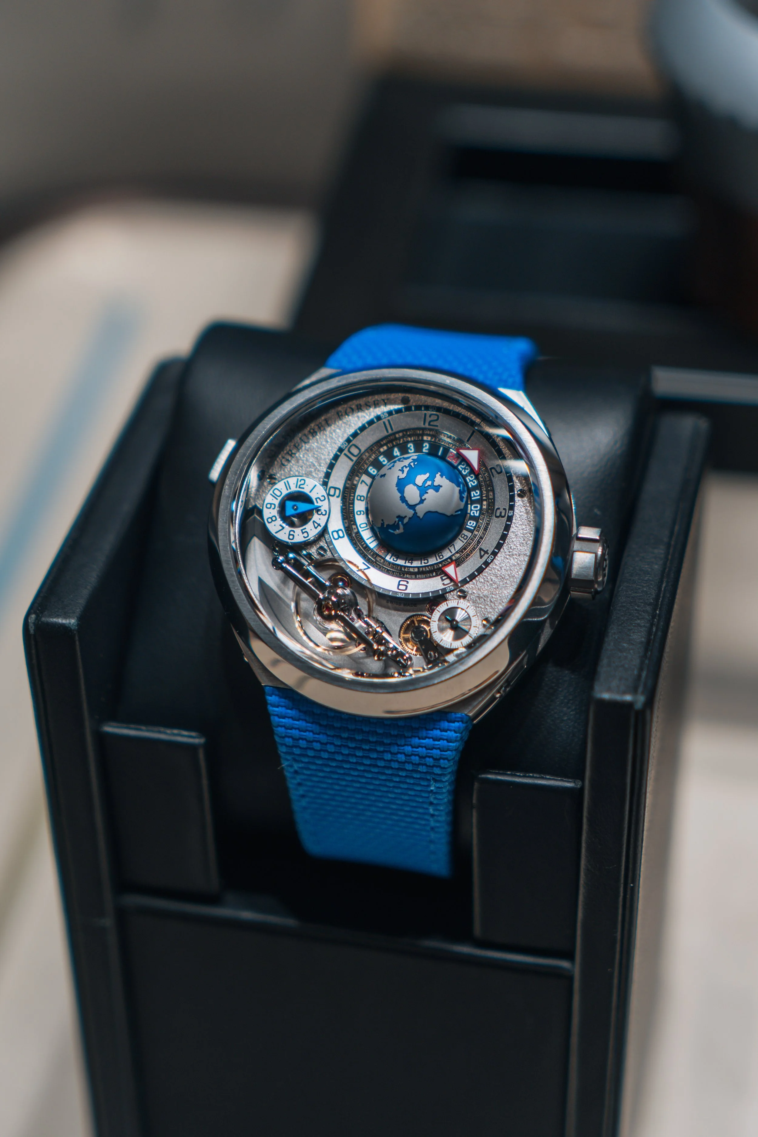 A luxury wristwatch with a blue strap, featuring a detailed dial with a globe, world time zones, and multiple dials, placed on a black display stand.