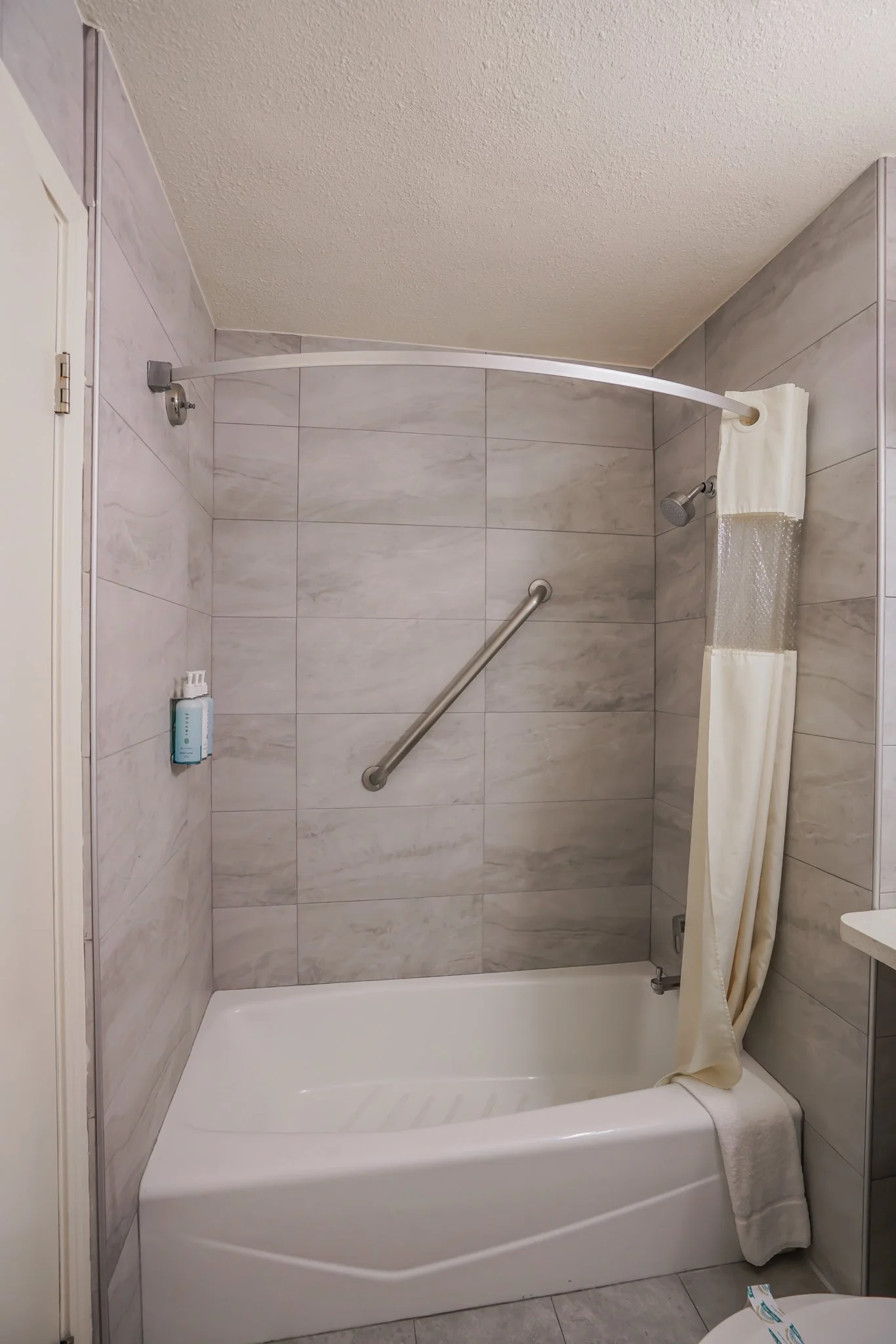 Bathroom shower with beige tile walls, a curved shower curtain rod with a partially open cream-colored shower curtain, a towel hanging on the shower, a grab bar, and a bottled soap or shampoo on a wall-mounted shelf.