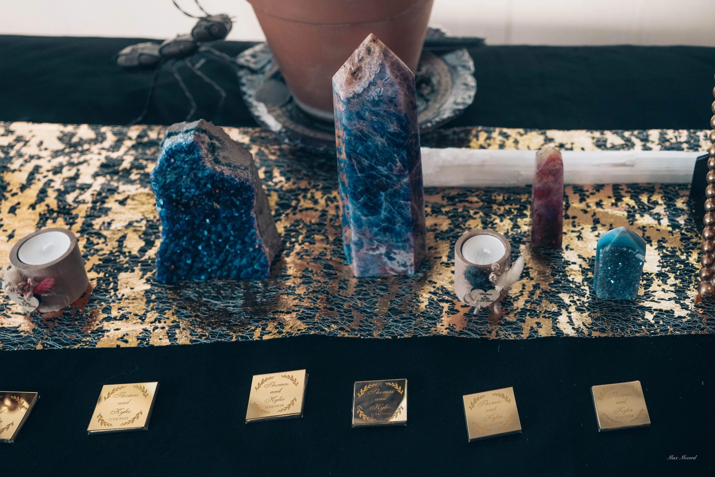 Decorative display with large blue and purple crystals, small candles, and small gold plaques with names and dates, set on a black and gold table runner.