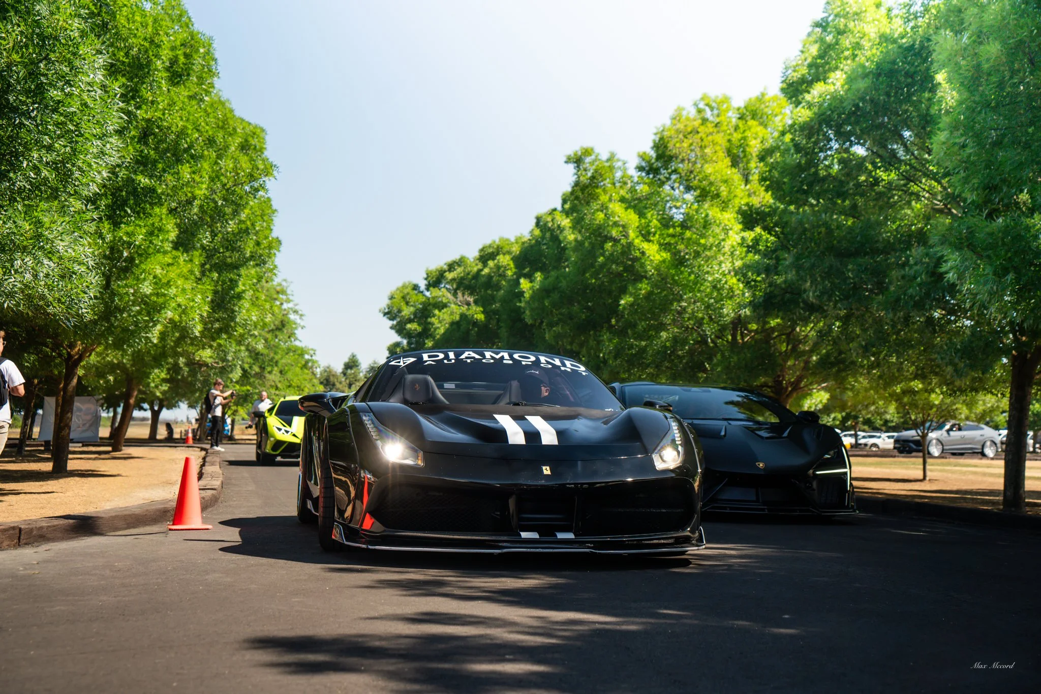 Black Ferrari sports car on a paved road surrounded by green trees, with a yellow Lamborghini in the background.