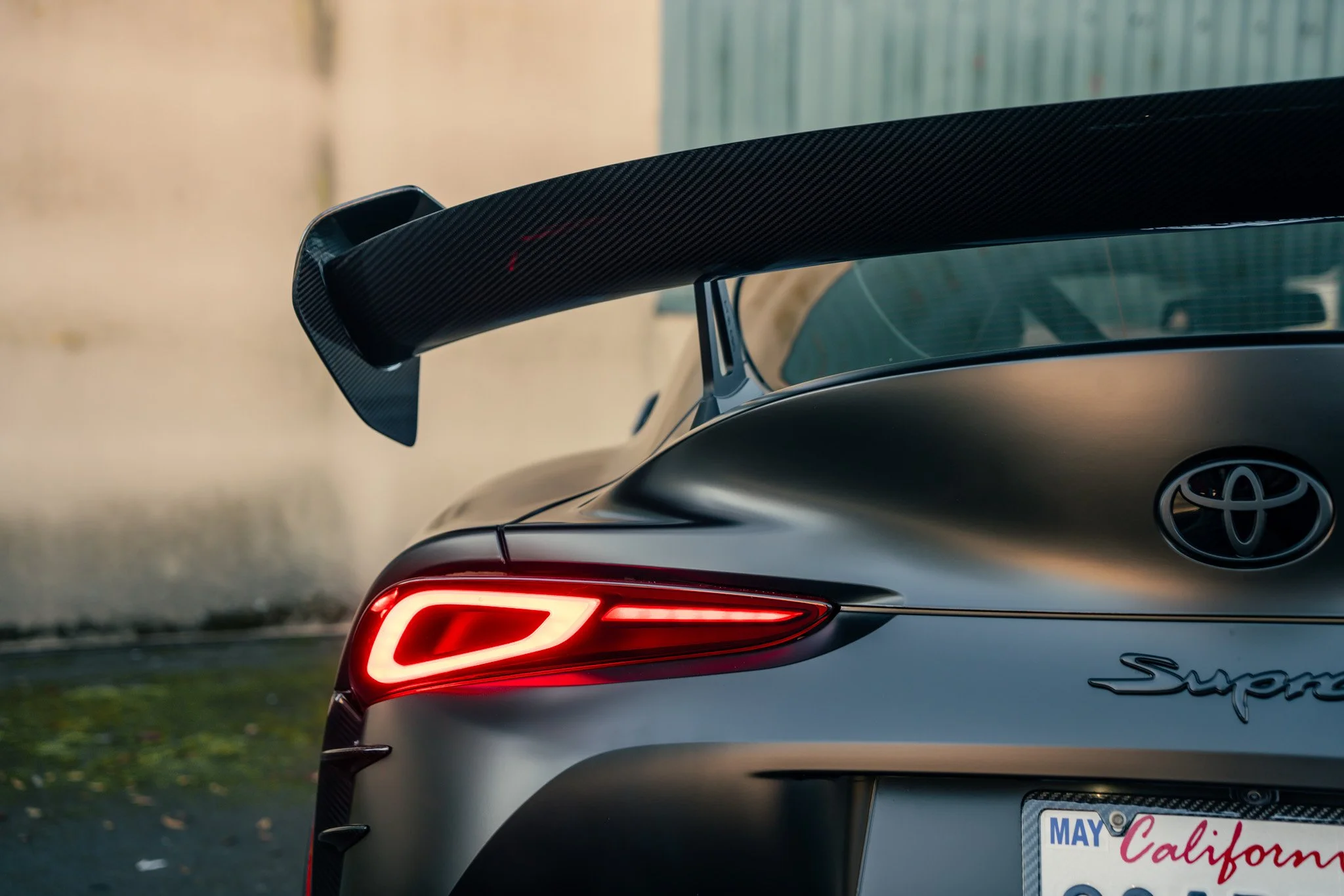 Close-up of the rear end of a matte gray Toyota Supra sports car with a black carbon fiber rear wing and illuminated tail light, parked outdoors.