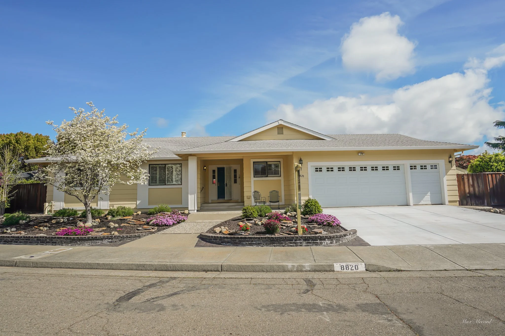 Front view of a beige single-story house with a gray roof, white garage door, and a small front porch. There is a blooming tree and flower beds with purple flowers in the yard. The house number 8820 is painted on the curb.