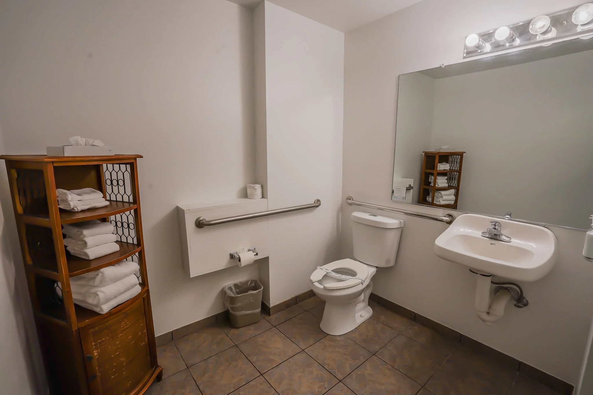 Accessible bathroom with toilet, wall-mounted sink, large mirror, towel rack, and wooden storage cabinet with towels.