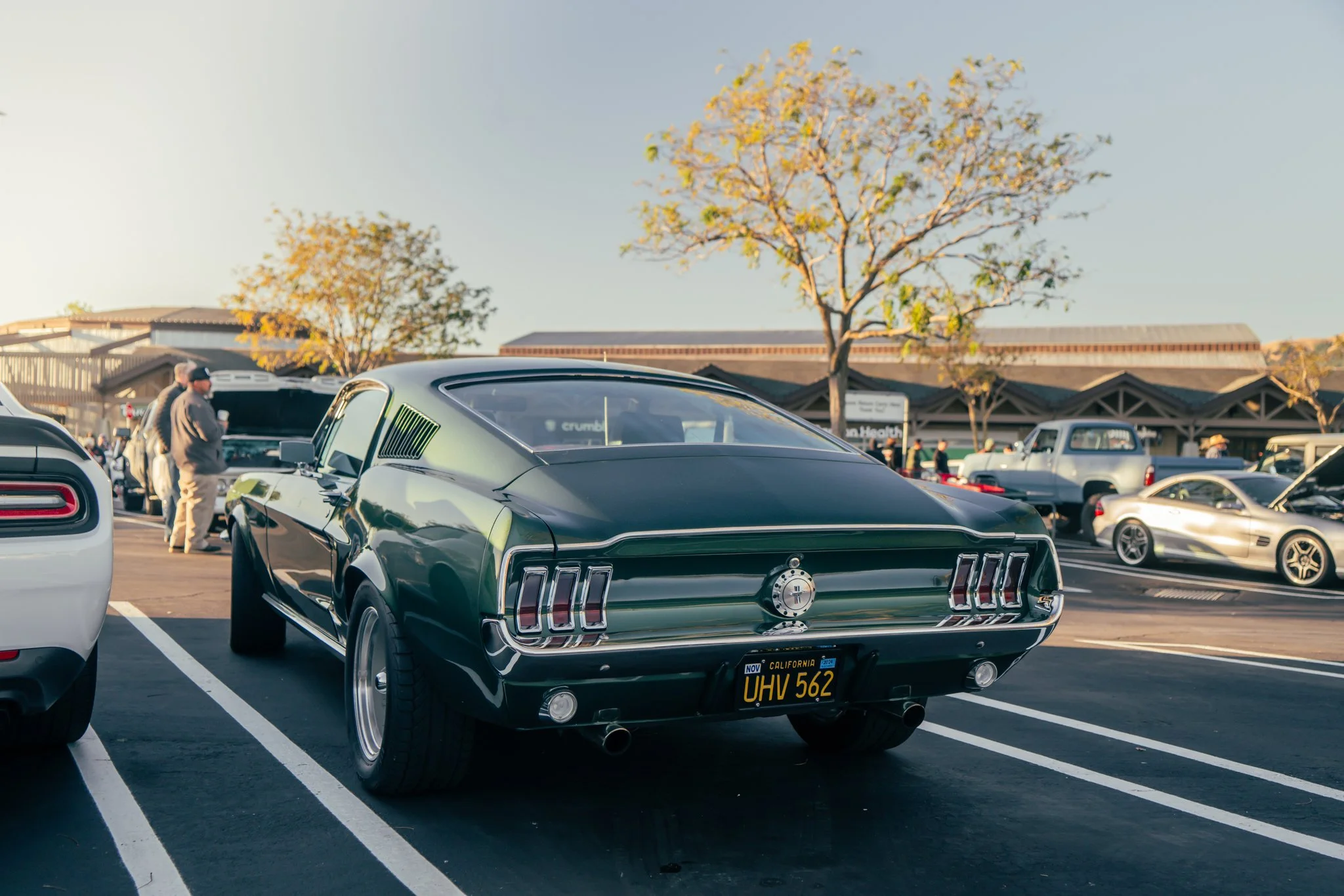 Rear view of a classic dark green Ford Mustang with a California license plate UHV 562 in a parking lot, surrounded by other cars and people at an outdoor car show.