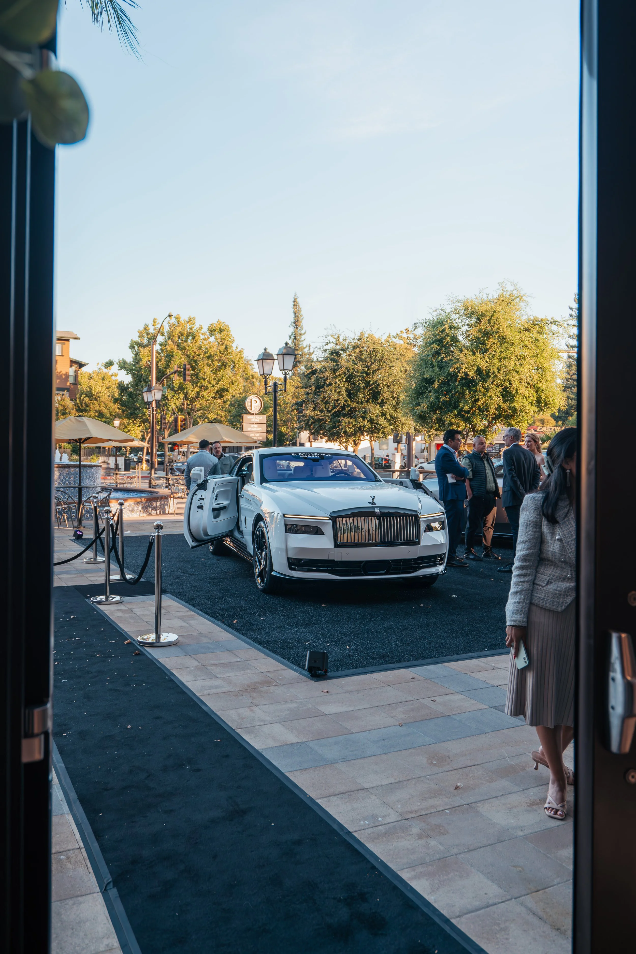 Luxury white car on display outside a building, with a woman in a business suit walking by and people engaging in conversation in the background, trees, and outdoor seating visible.
