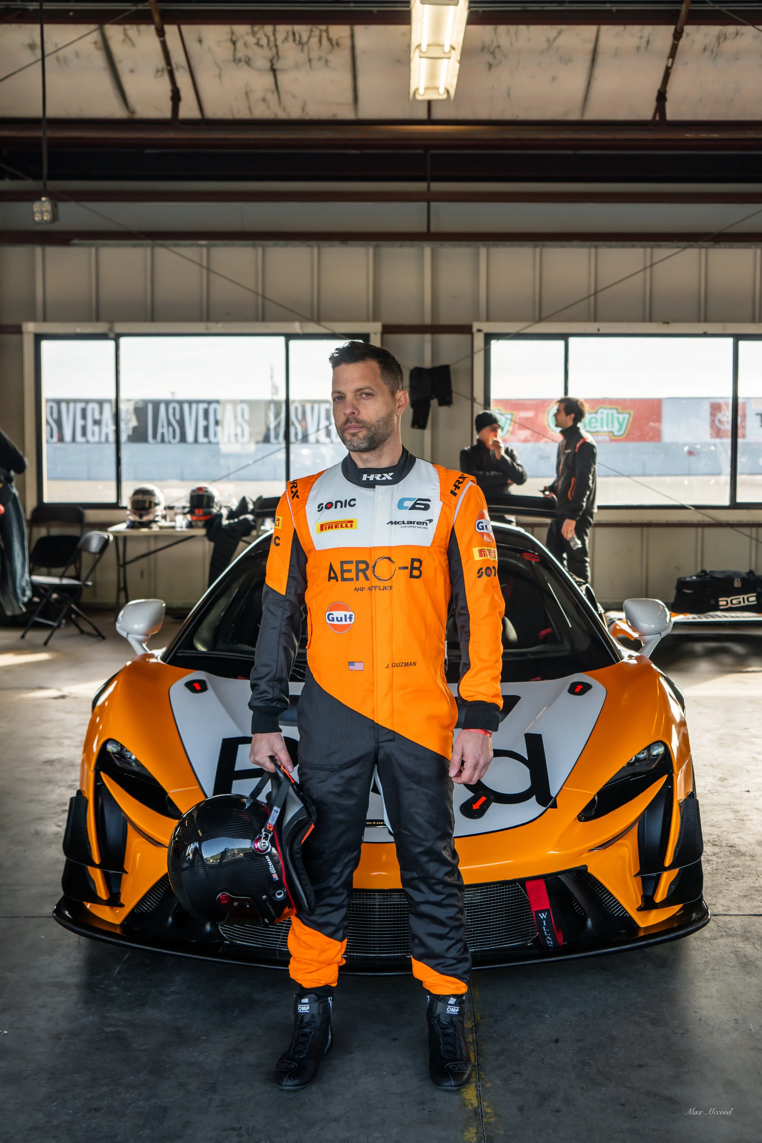 Race car driver in black and orange racing suit with helmet, standing in garage with racing car and other team members in background.