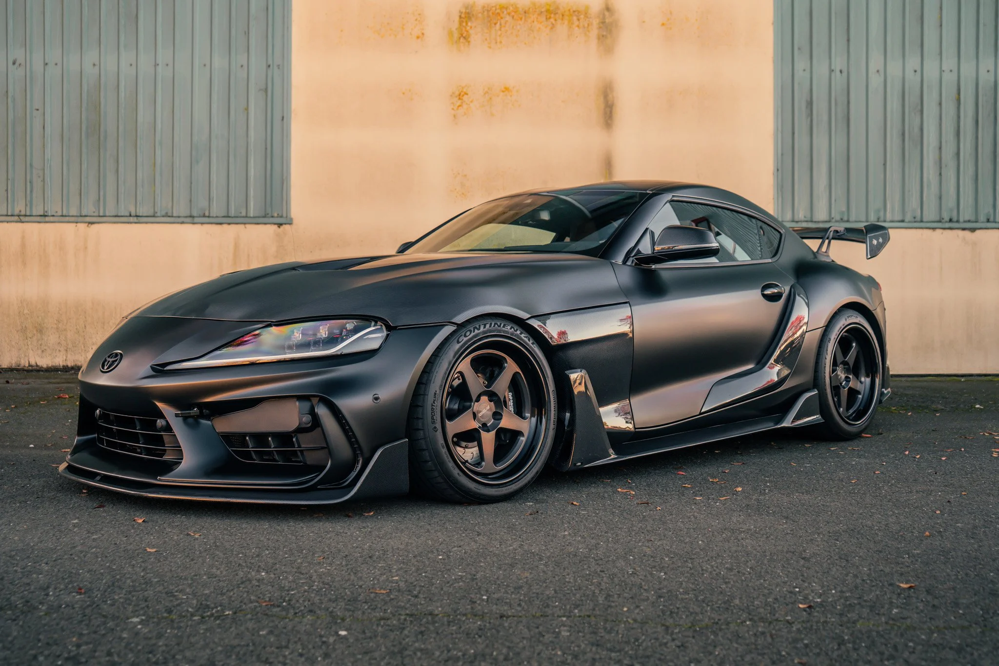 A matte black sports car parked on a paved surface in front of a building with metal siding.