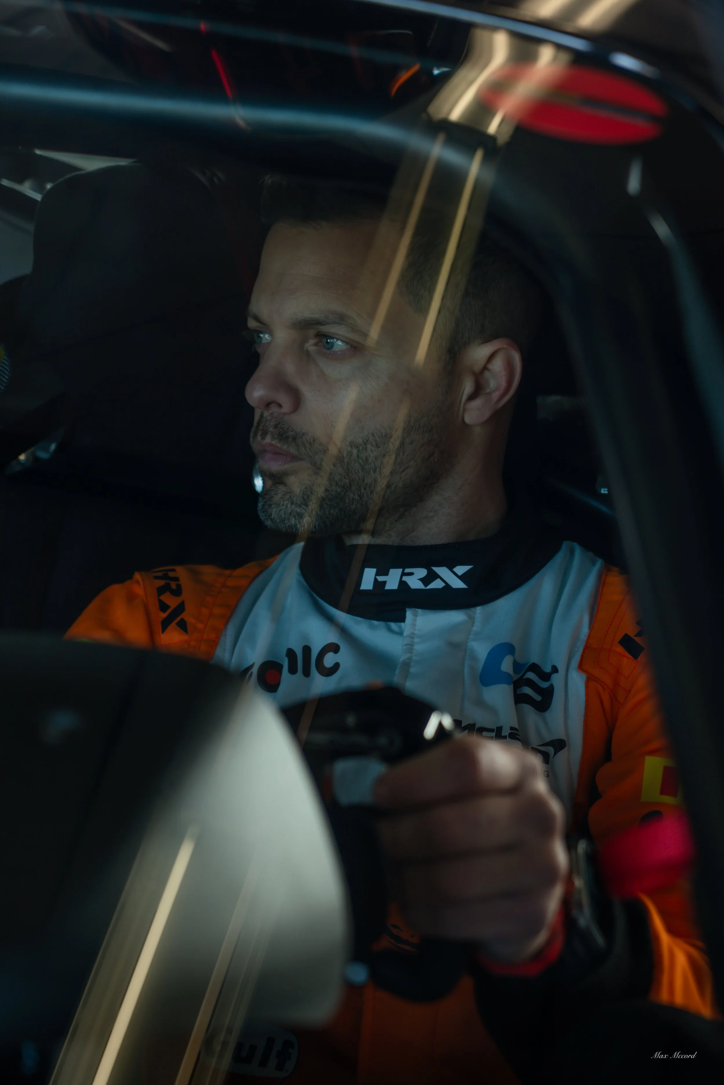 A male race car driver sitting inside his vehicle, wearing a racing suit and holding a steering wheel.