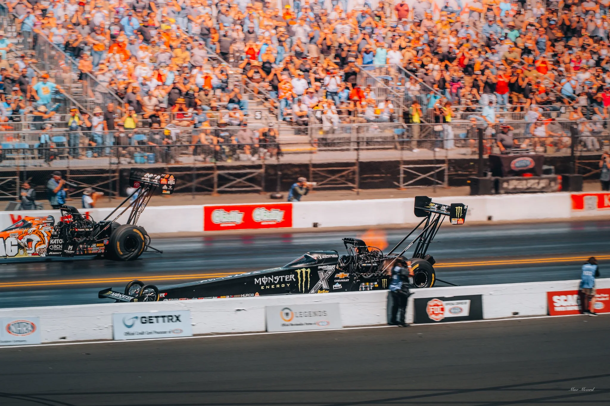 Two Monster Energy drag race cars on a racetrack preparing for a race, with a large crowd in the stands watching.