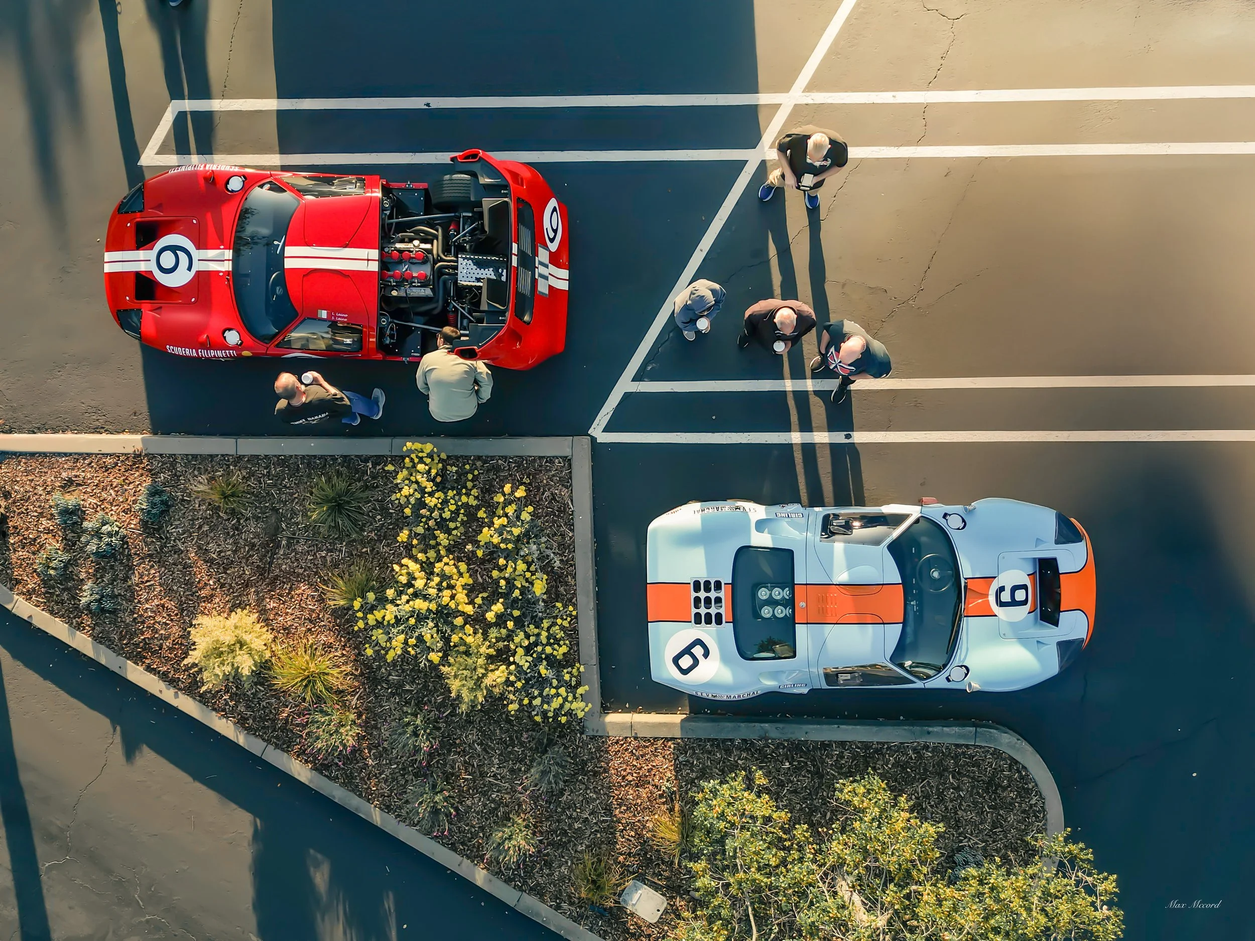 An aerial view of a parking lot with two vintage race cars, one red and one white with orange stripes, and four people standing nearby, some inspecting the red car.