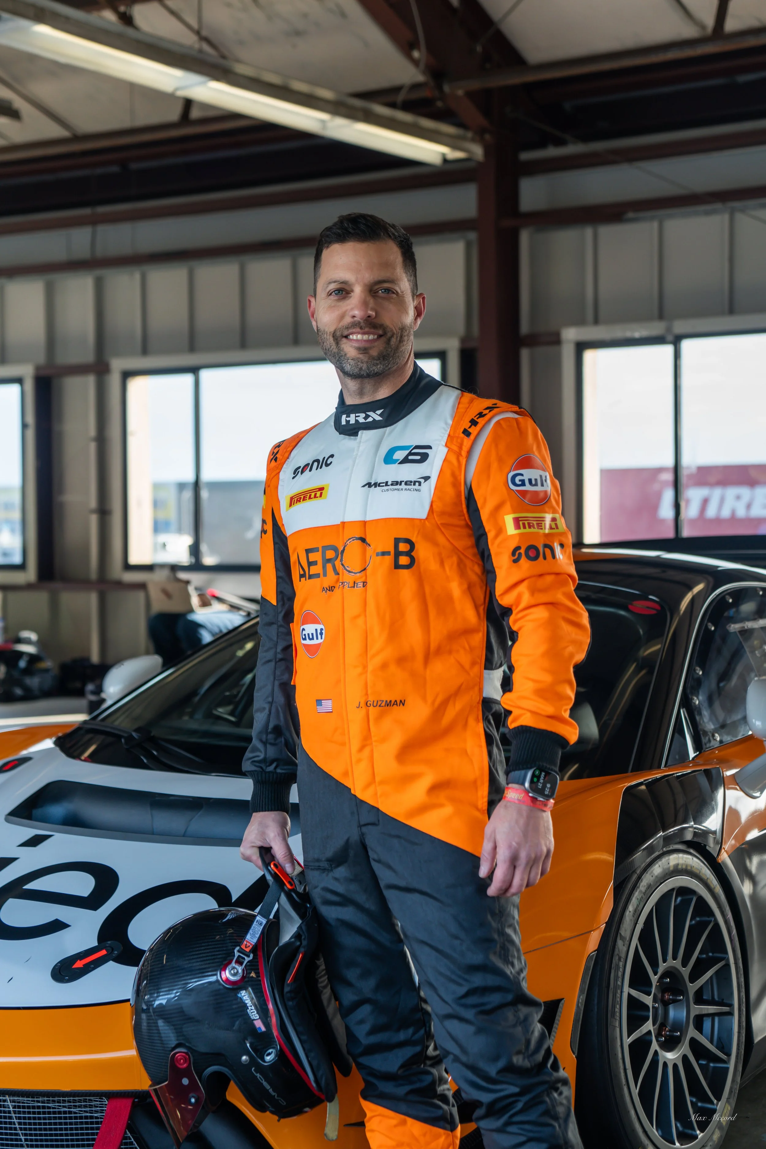 A race car driver standing in a garage with a racing helmet in hand, wearing an orange and black racing suit.
