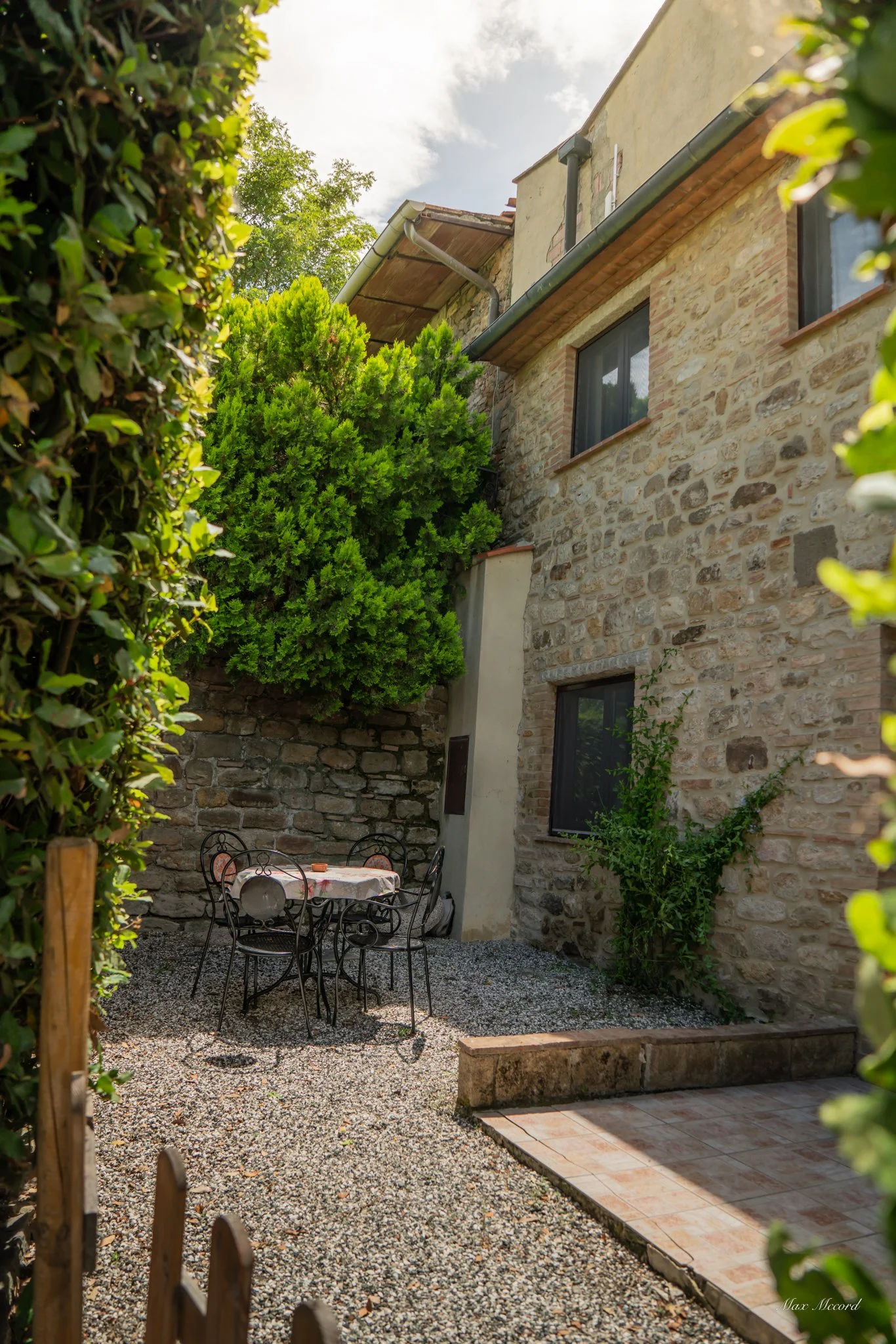 Small outdoor patio with a round table covered with a pink tablecloth, four black wrought iron chairs, surrounded by stone walls, green trees, and bushes under a partly cloudy sky.