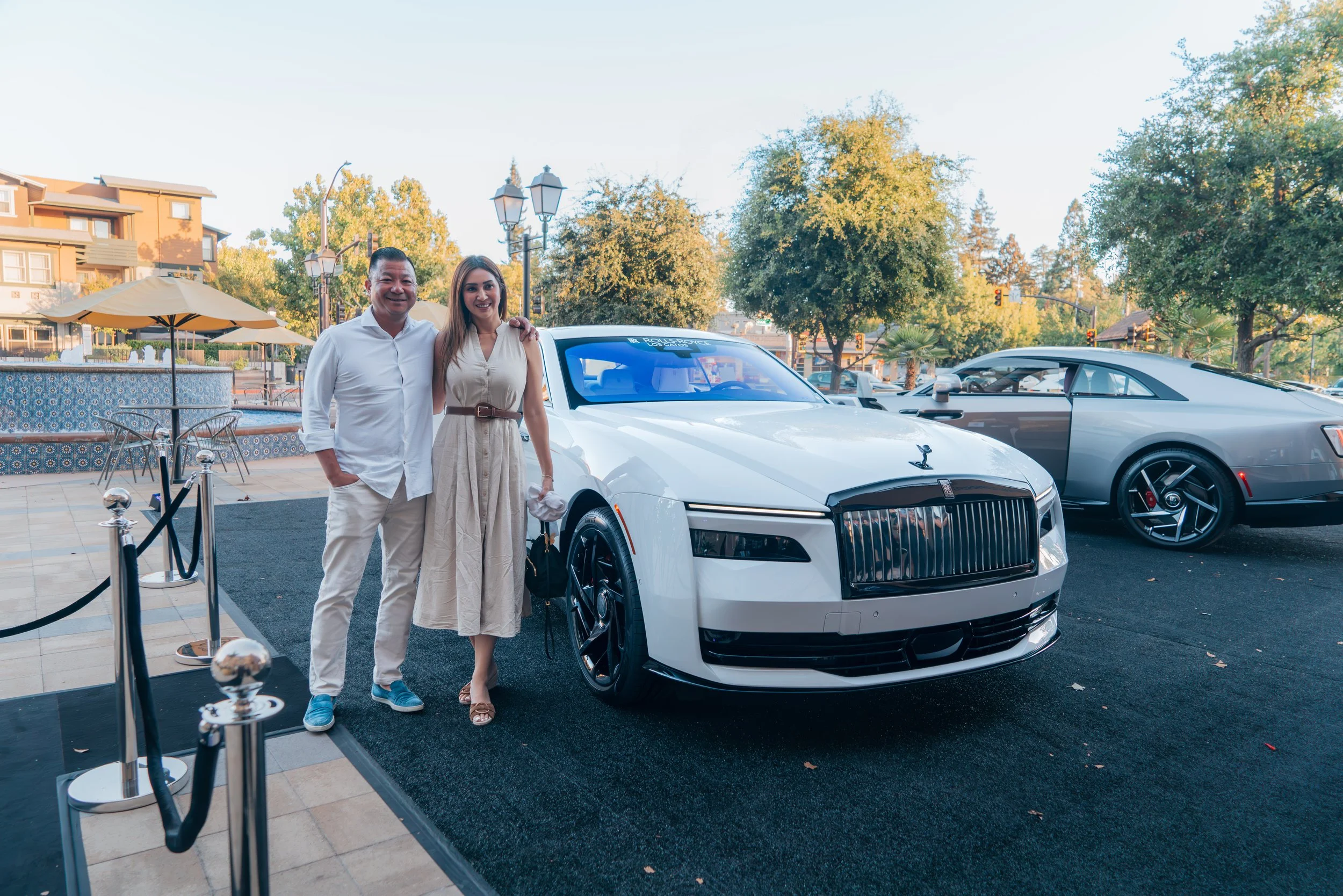 Two people standing next to a white luxury car at an outdoor event with trees and additional cars in the background.