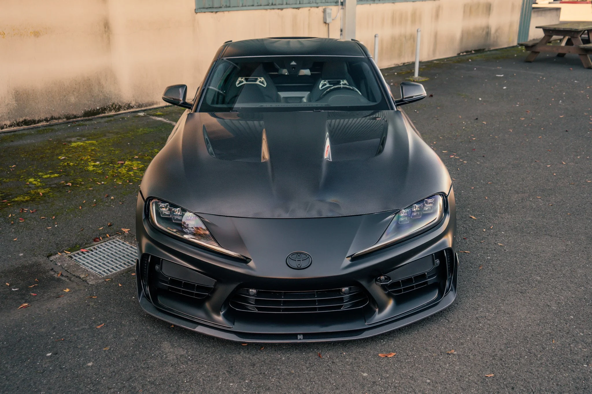 Front view of a black Toyota sports car with aerodynamic design and hood vents, parked on asphalt near a beige wall.