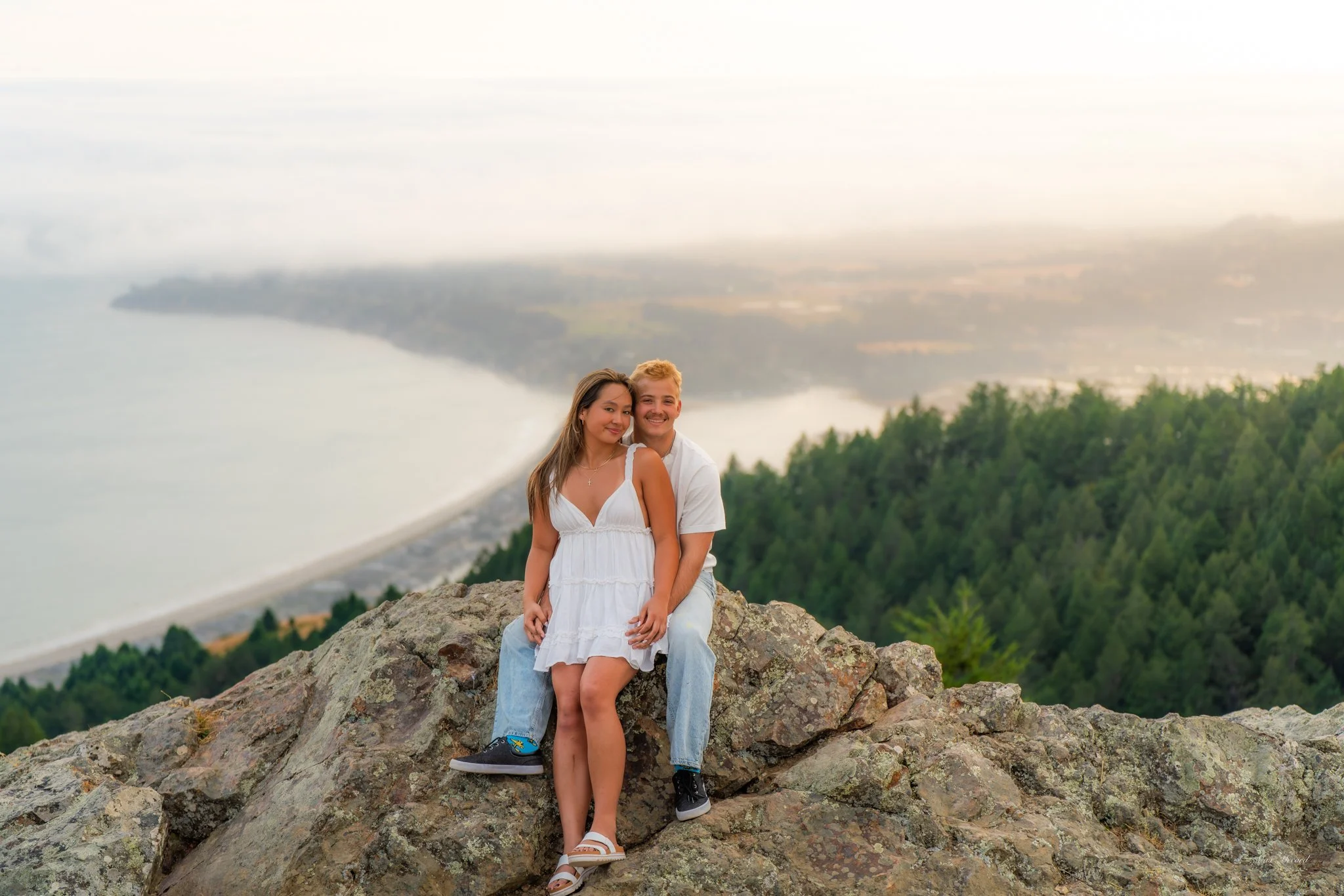 A young couple sitting on a rocky ledge overlooking a scenic view of a coastline with the ocean, forested hills, and a distant shoreline.