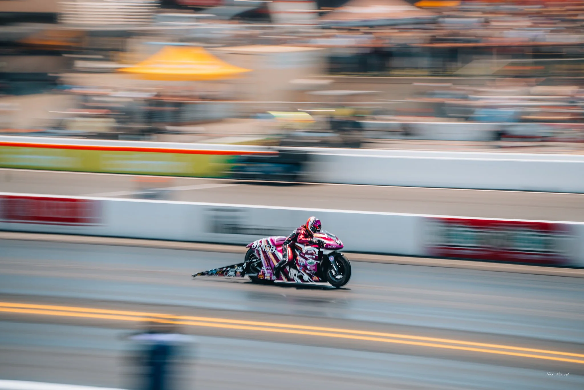 Motorsport motorcycle racer on a racing track, leaning forward on a pink and white motorcycle, with blurred background and track surroundings, indicating high speed.