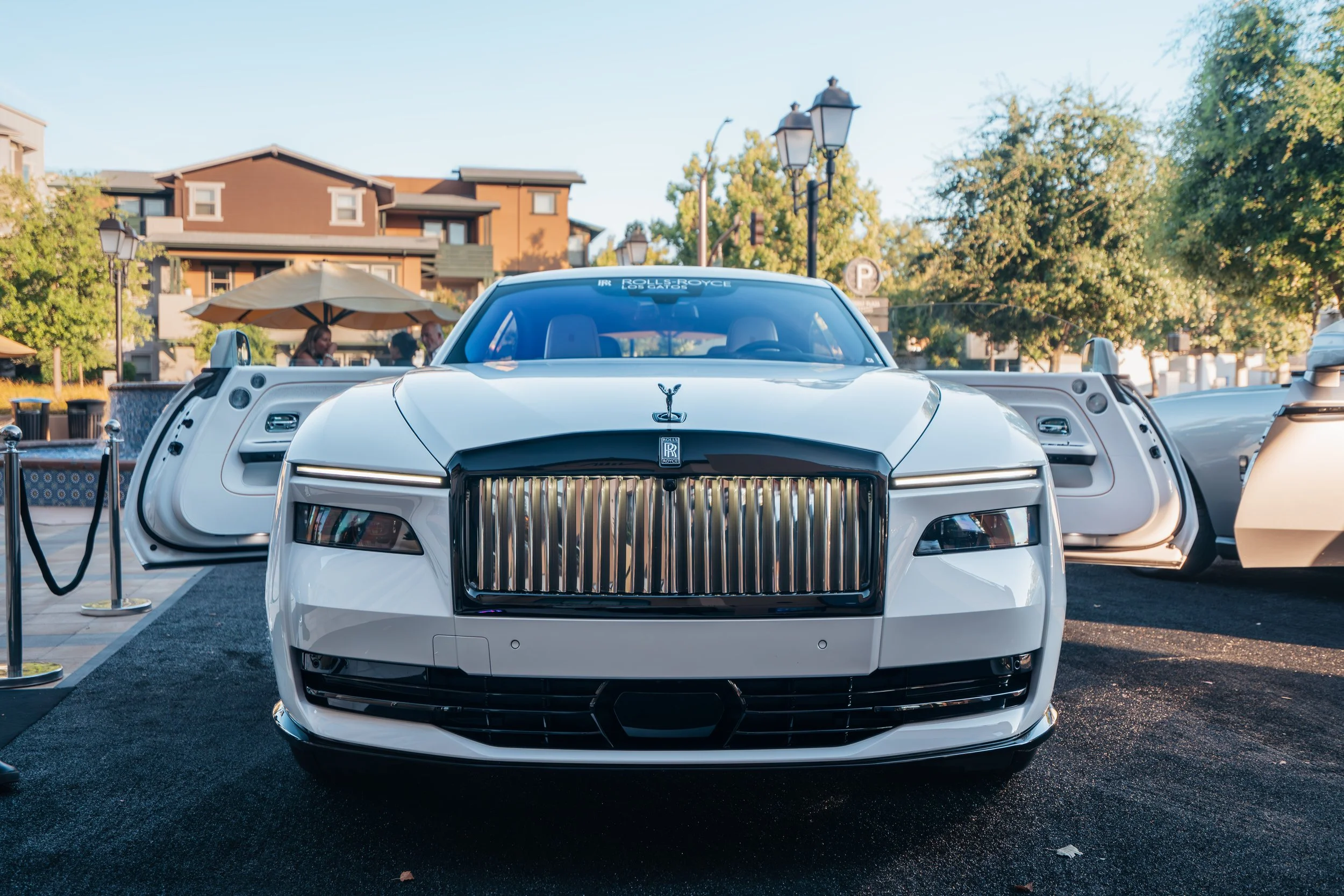White luxury Rolls-Royce car with a large chrome grille on display at an outdoor event, with open doors and a backdrop of trees, buildings, and people.