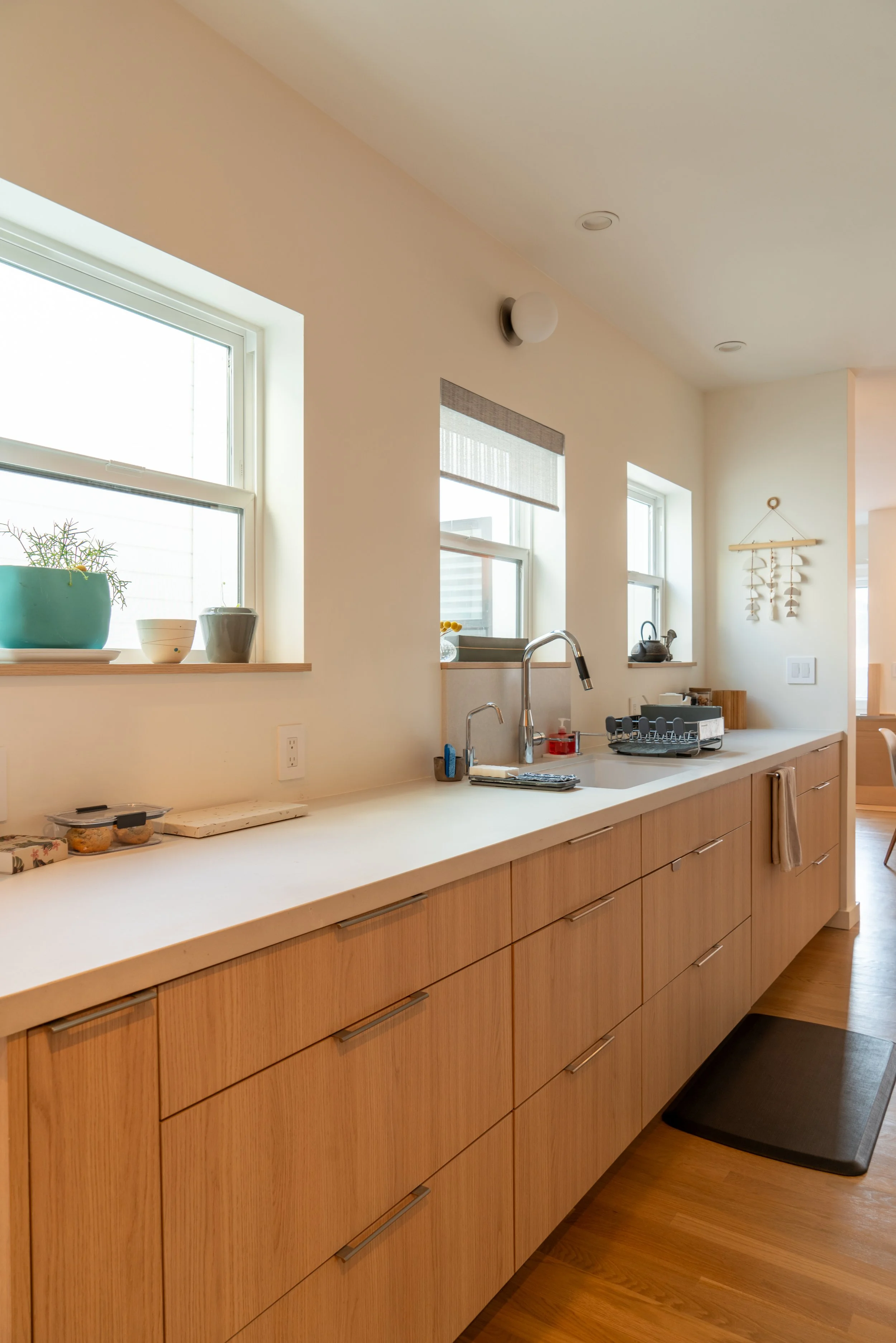 Modern kitchen with light wood cabinets, white countertop, three small windows with blinds, and decor on the windowsills.
