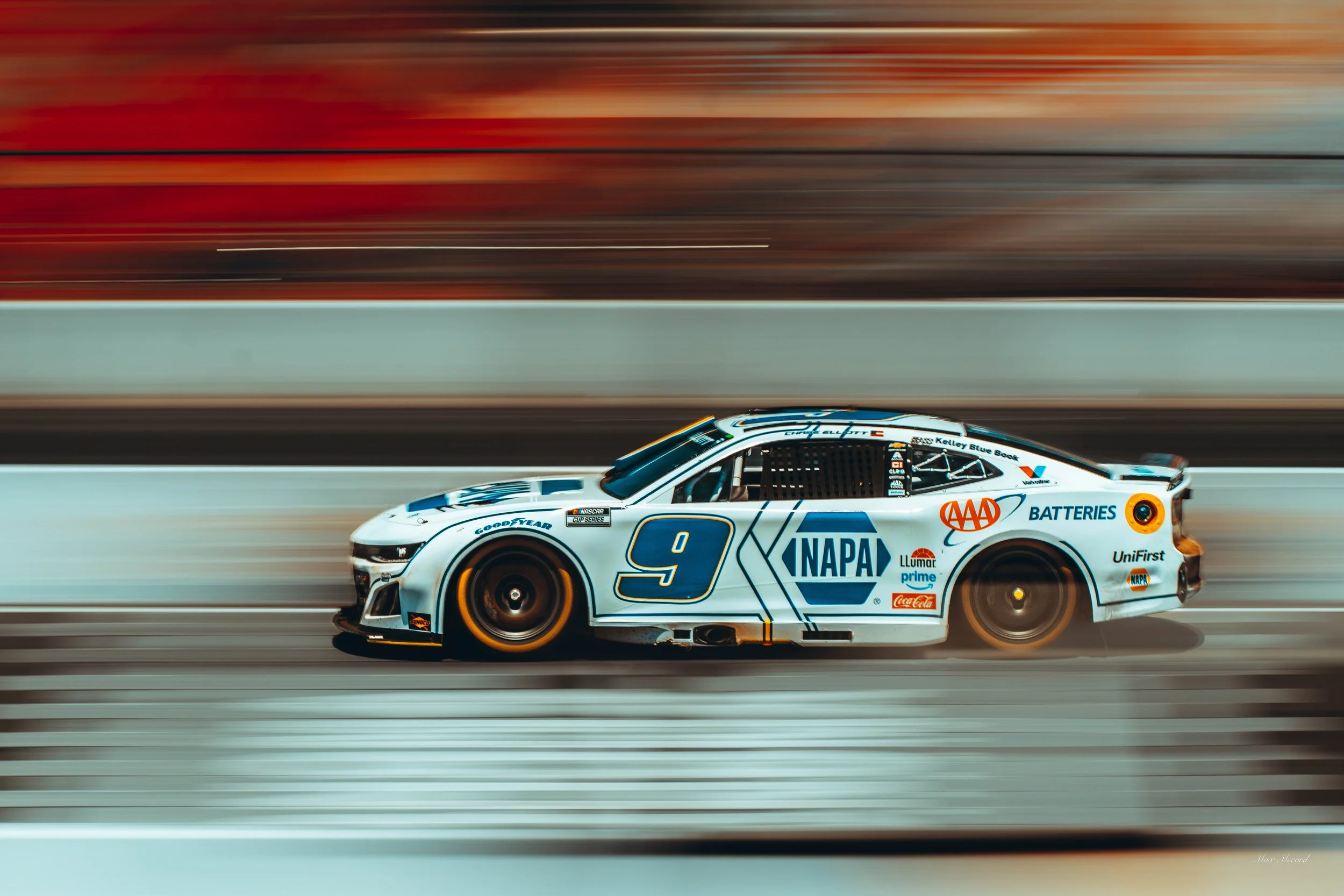 Race car with sponsor logos, number 9, moving at high speed on a racetrack with blurred background.