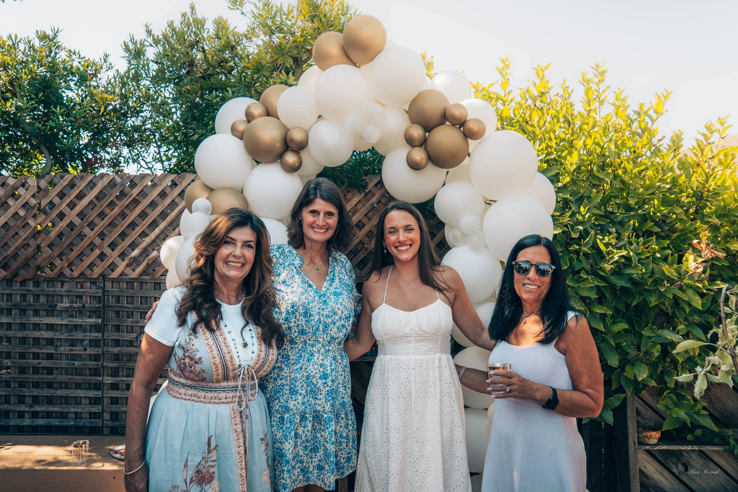 Four women standing together at an outdoor celebration in front of a balloon arch and a wooden fence with green trees, sunny weather.