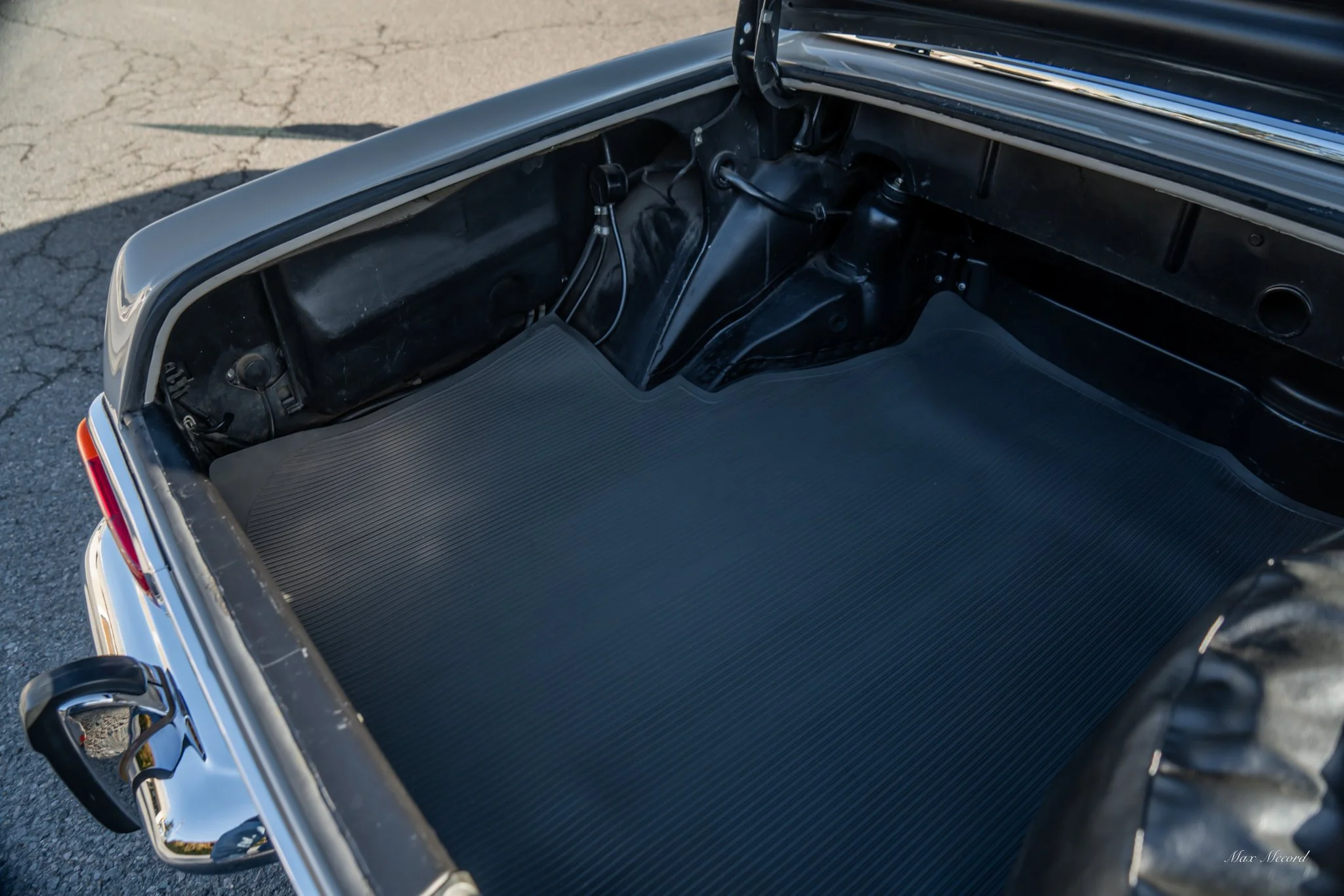 Empty vintage car trunk with a black interior and textured floor, parked on pavement.