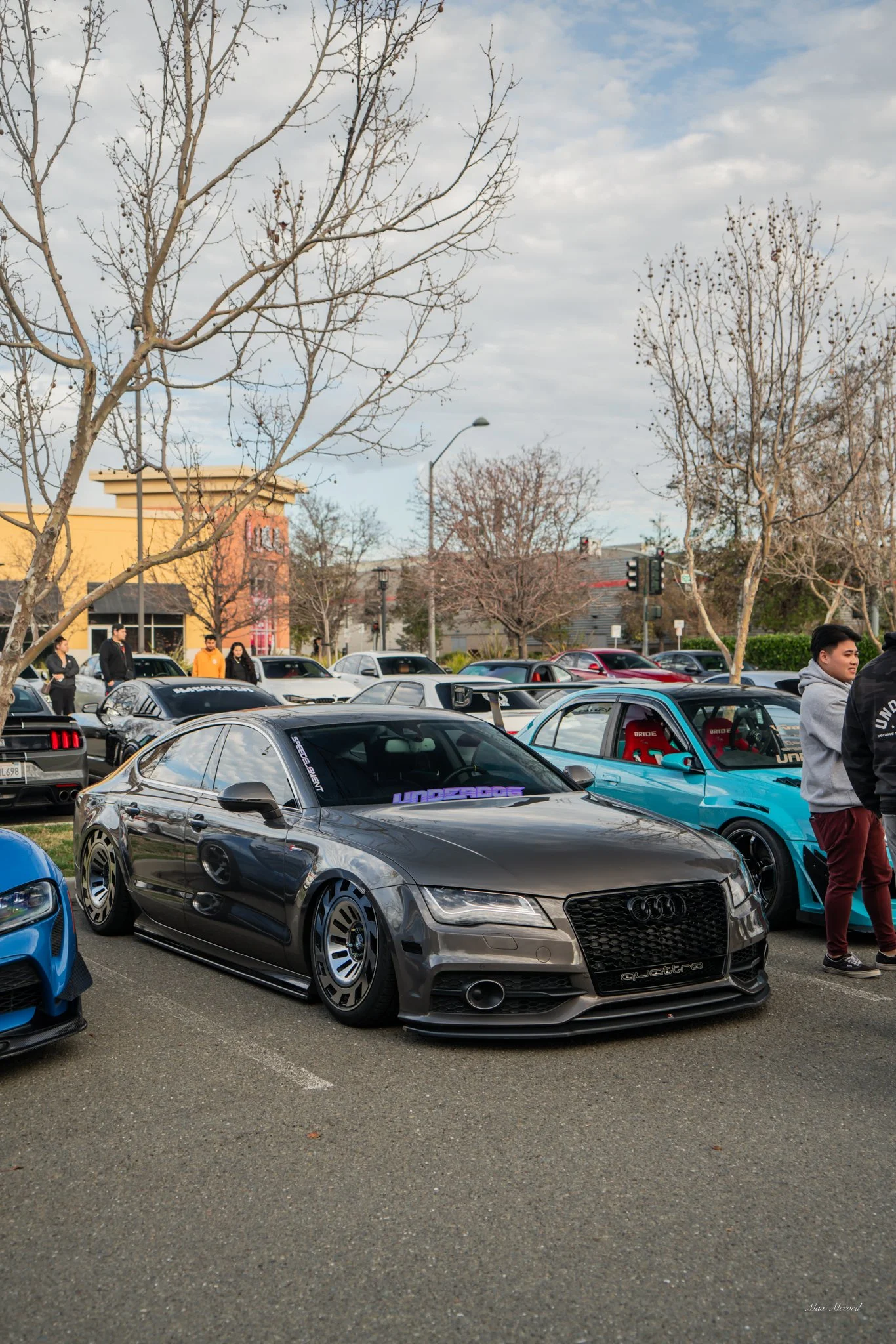 Black Audi parked at a car show, surrounded by other modified cars and people, with leafless trees and a partly cloudy sky in the background.