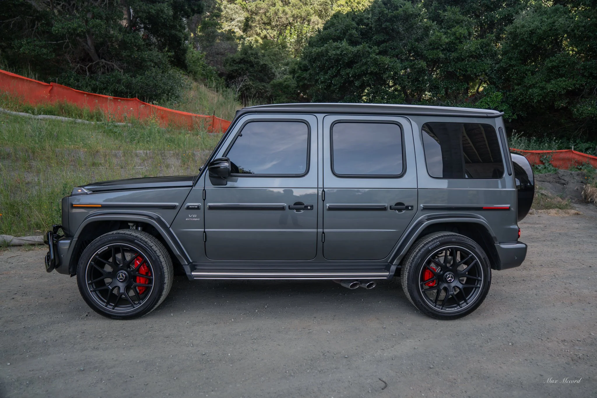 A gray Mercedes-Benz G-Class SUV parked on a dirt road near greenery and a hillside.