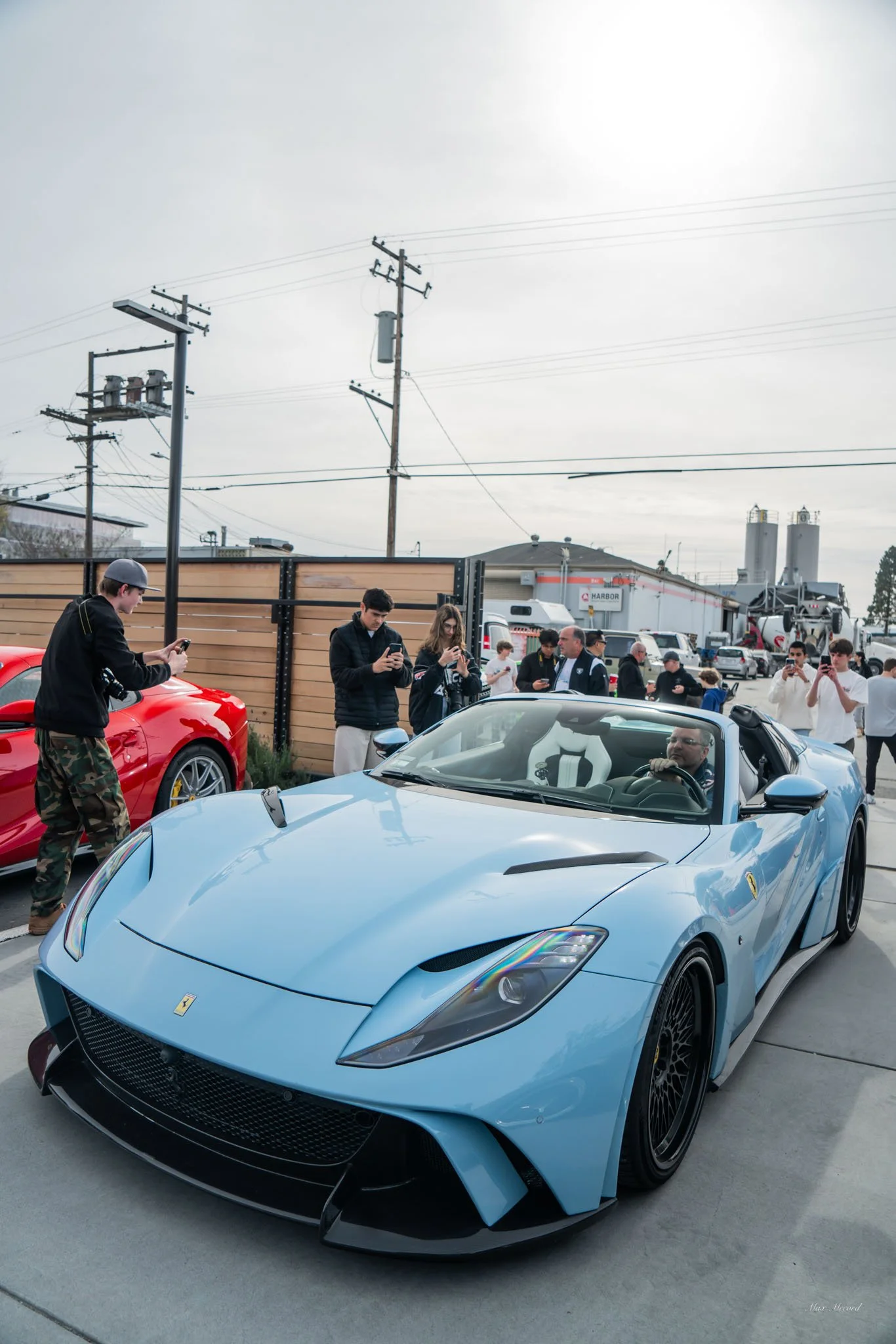 A light blue Ferrari convertible sports car parked outdoors with several people around it, some taking photos and others observing. The background includes power lines and industrial buildings.