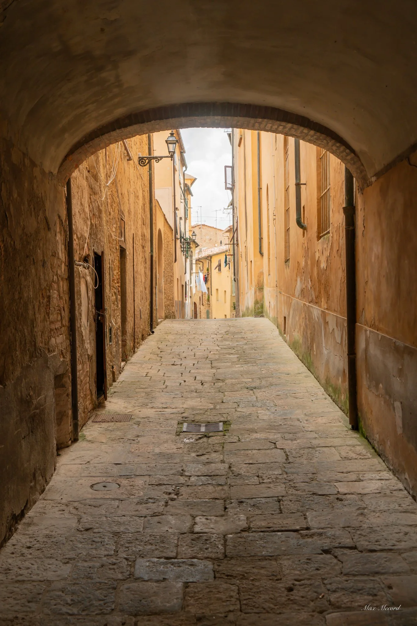Narrow cobblestone alleyway in a historic European town, viewed from under an archway, with warm yellow and brown buildings on either side and a glimpse of the sky in the distance.