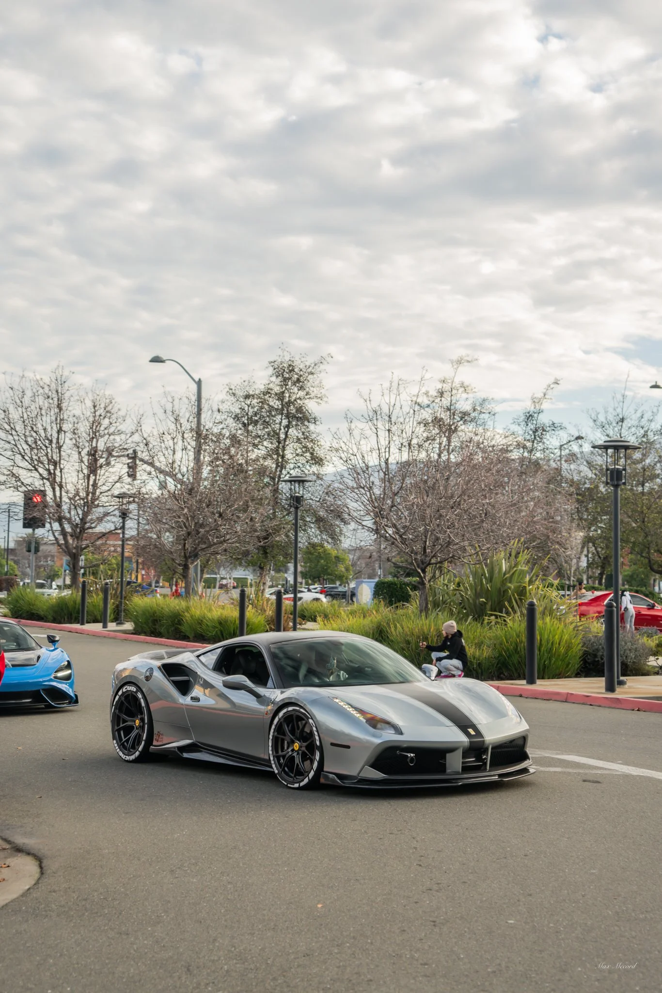 A silver Ferrari sports car parked on a city street with another blue sports car partially visible to the left. A person is sitting on a ledge behind the silver car, taking a photo or looking at their phone, with leafless trees, street lamps, and som