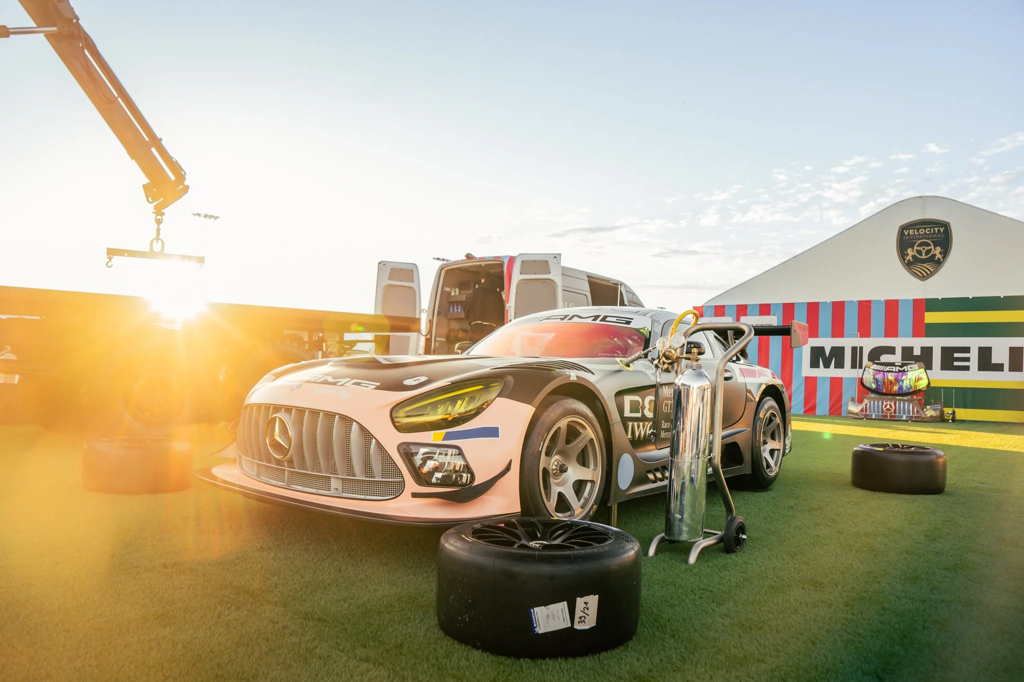 Race car in a pit area with a tire, oxygen tank, and tools, with a tent bearing the Michelin logo and a Velocify sign in the background, during sunset.