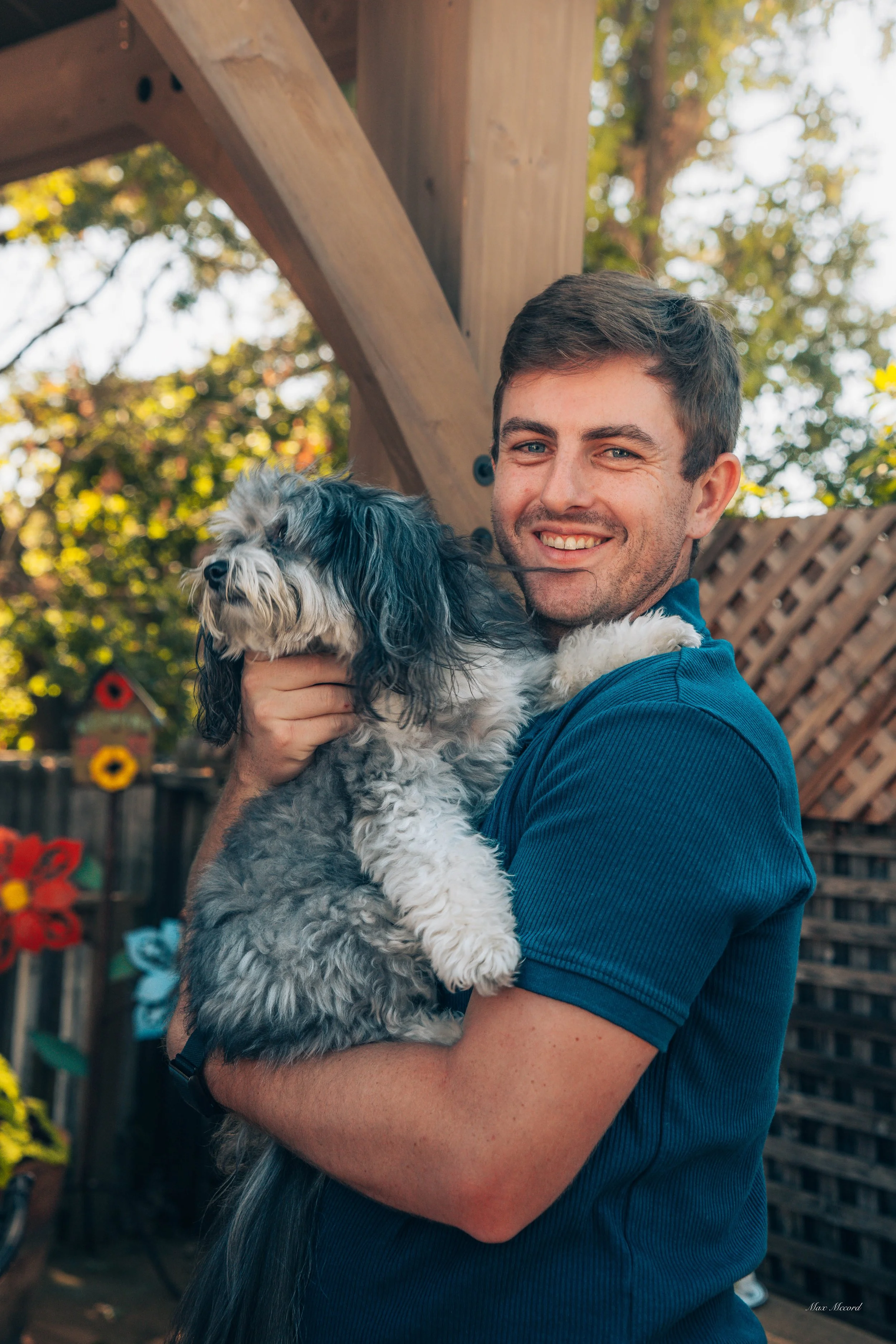 A smiling man in a blue shirt holding a fluffy black and white dog outdoors, with trees and a wooden fence in the background.