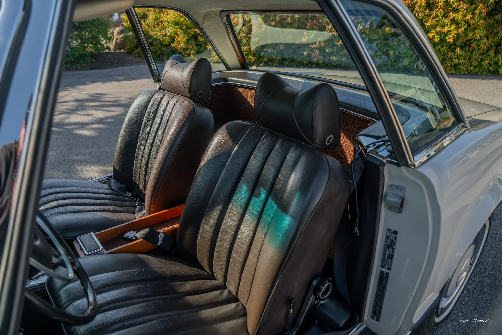 The interior of a classic car showing black leather seats, black headrests, and a wooden center console, with a view through the windshield of greenery outside.