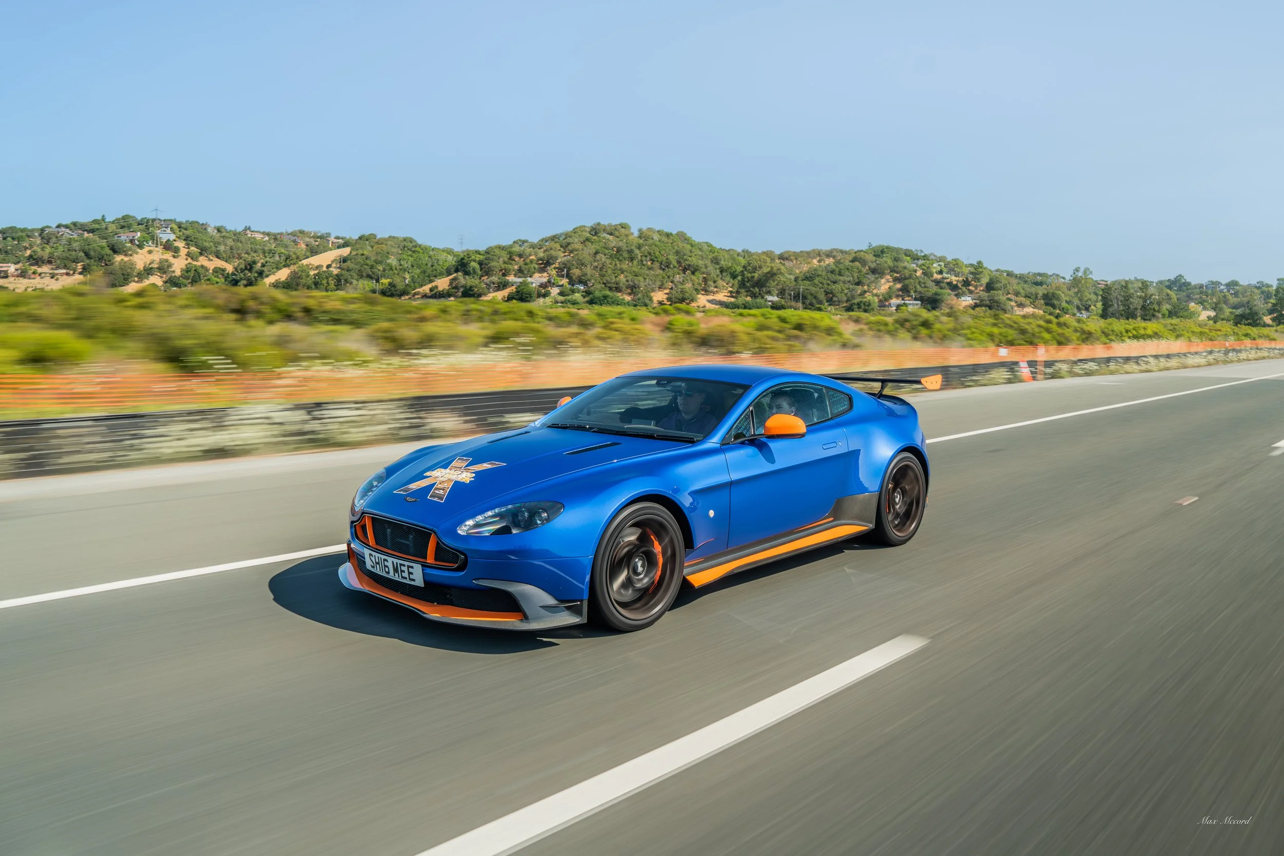 A blue race car with orange accents driving on a highway with a landscape of hills and trees in the background.