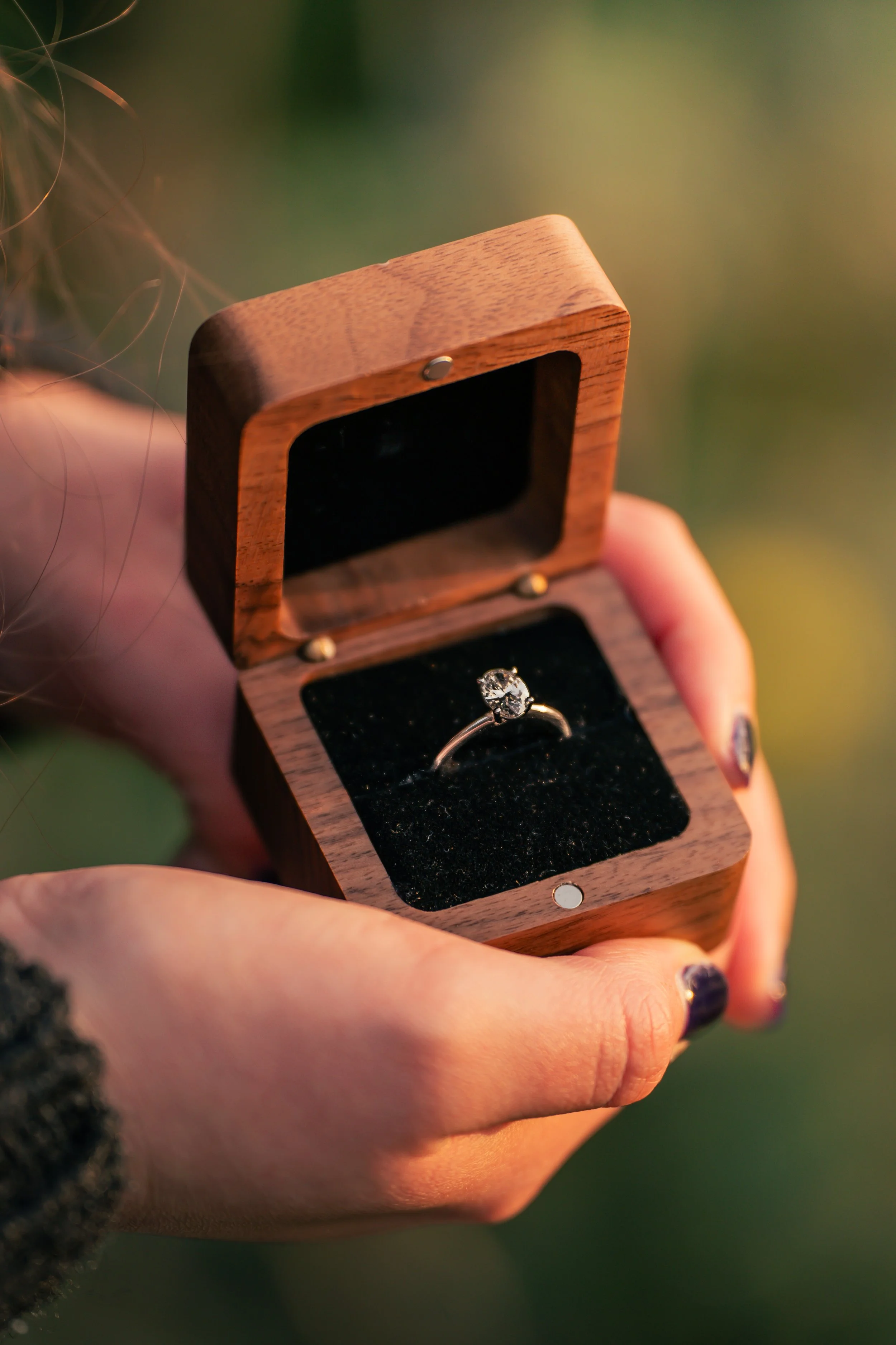 A person holding an open wooden ring box with a diamond engagement ring inside.