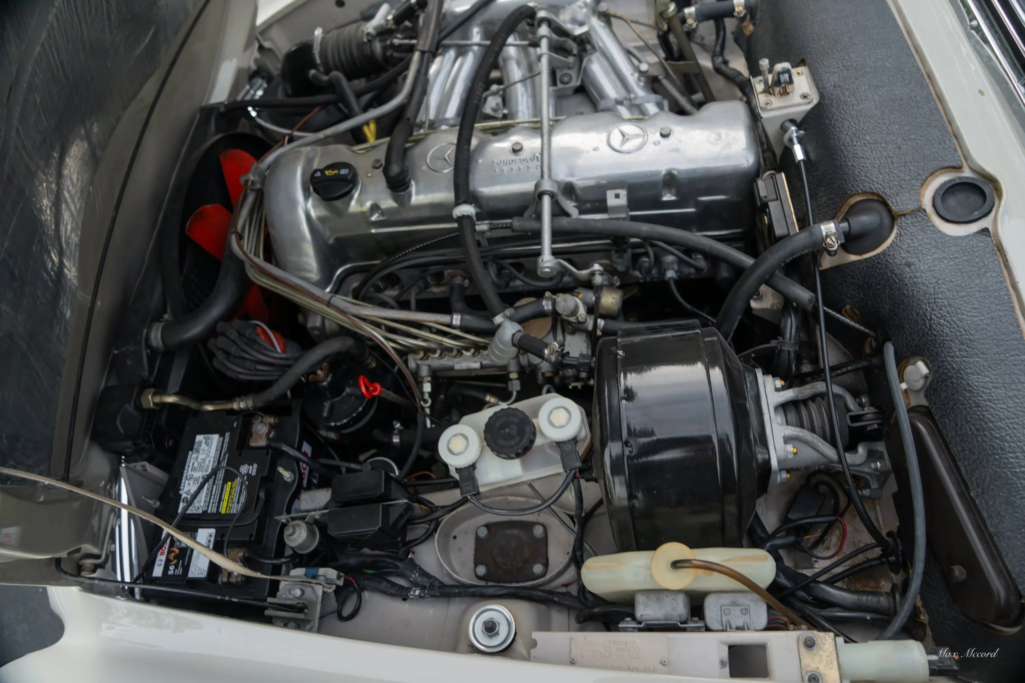 Engine bay of a classic Mercedes-Benz with visible engine components, black hoses, an aluminum valve cover, a black air filter box, and a vehicle battery.