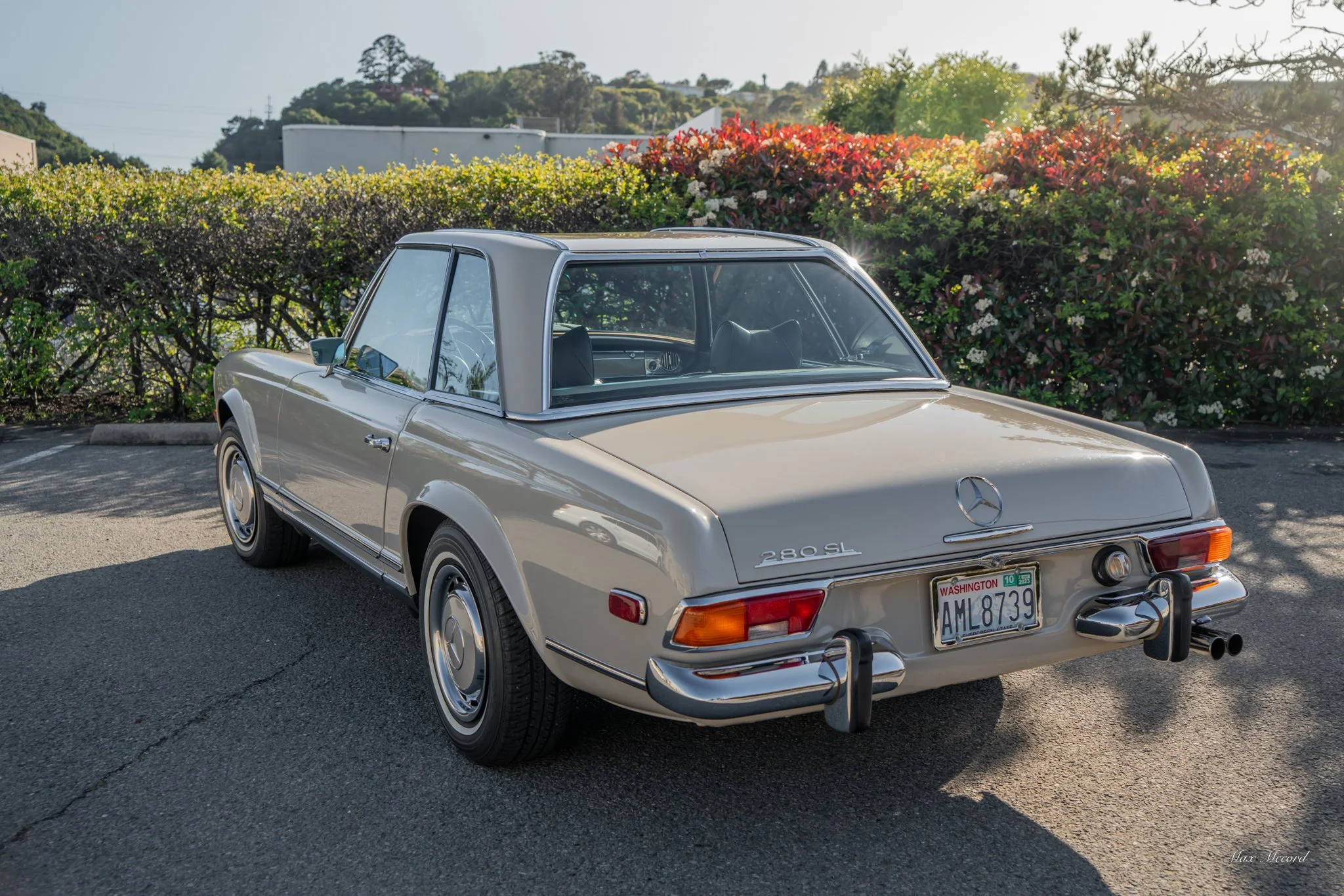 A vintage cream-colored Mercedes-Benz 280SL convertible car parked on an asphalt surface with greenery and bushes in the background.