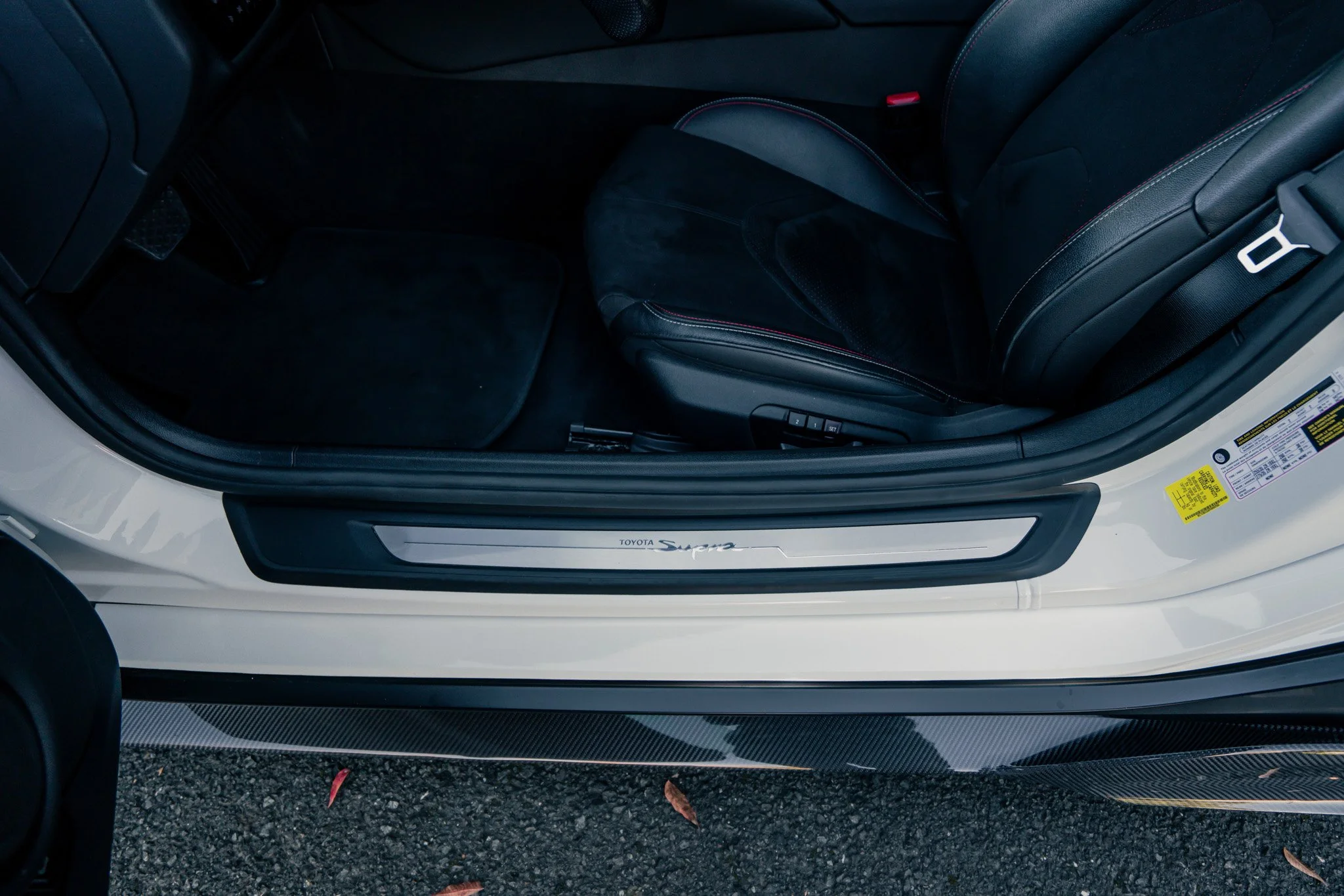 Close-up of the interior of a Toyota Supra, showing the black seat, door sill with 'TOYOTA Supra' logo, and part of the floor on a textured surface with fallen leaves.
