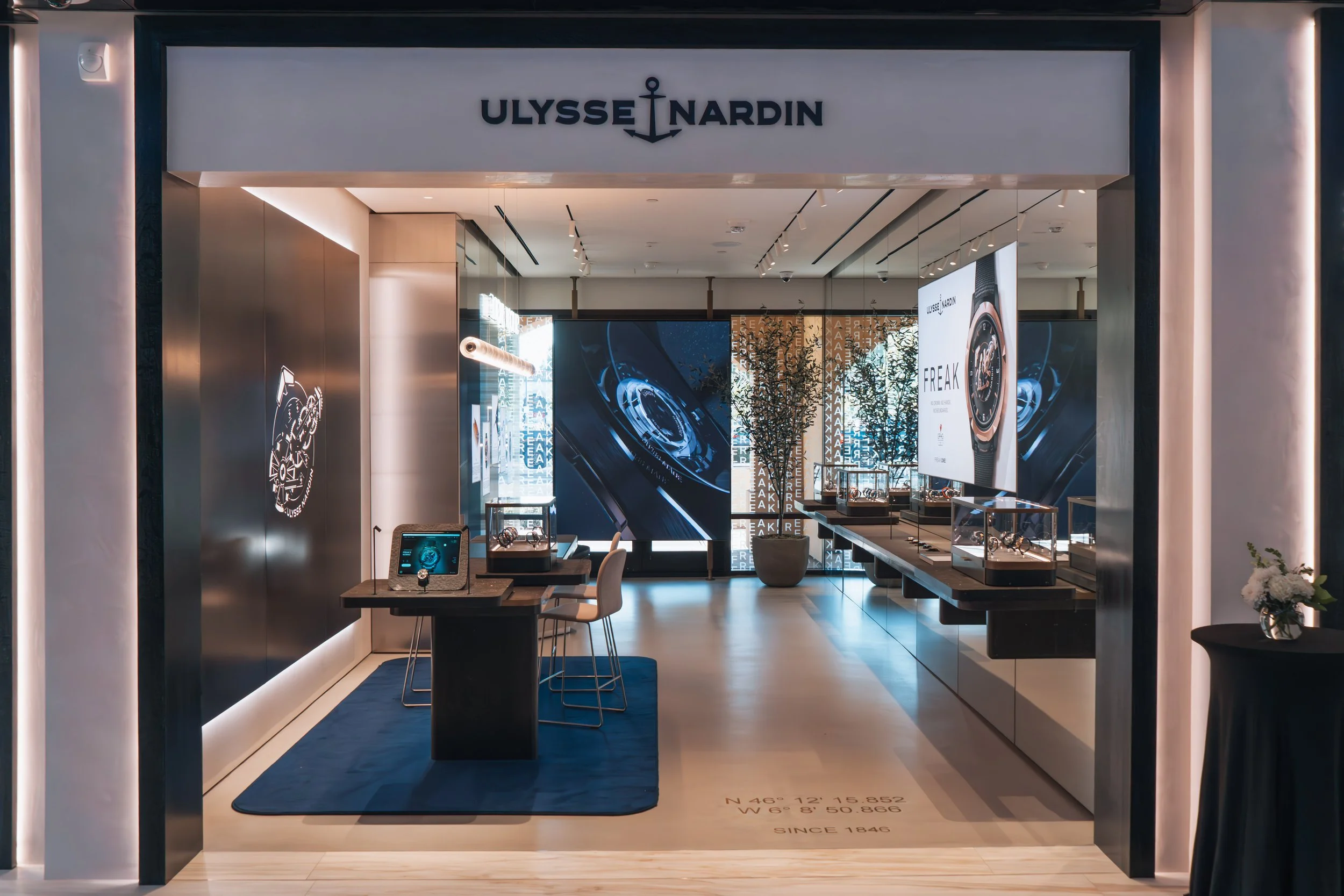 Ulysse Nardin boutique interior showcasing luxury watches, digital displays, and modern decor with trees and a blue rug.