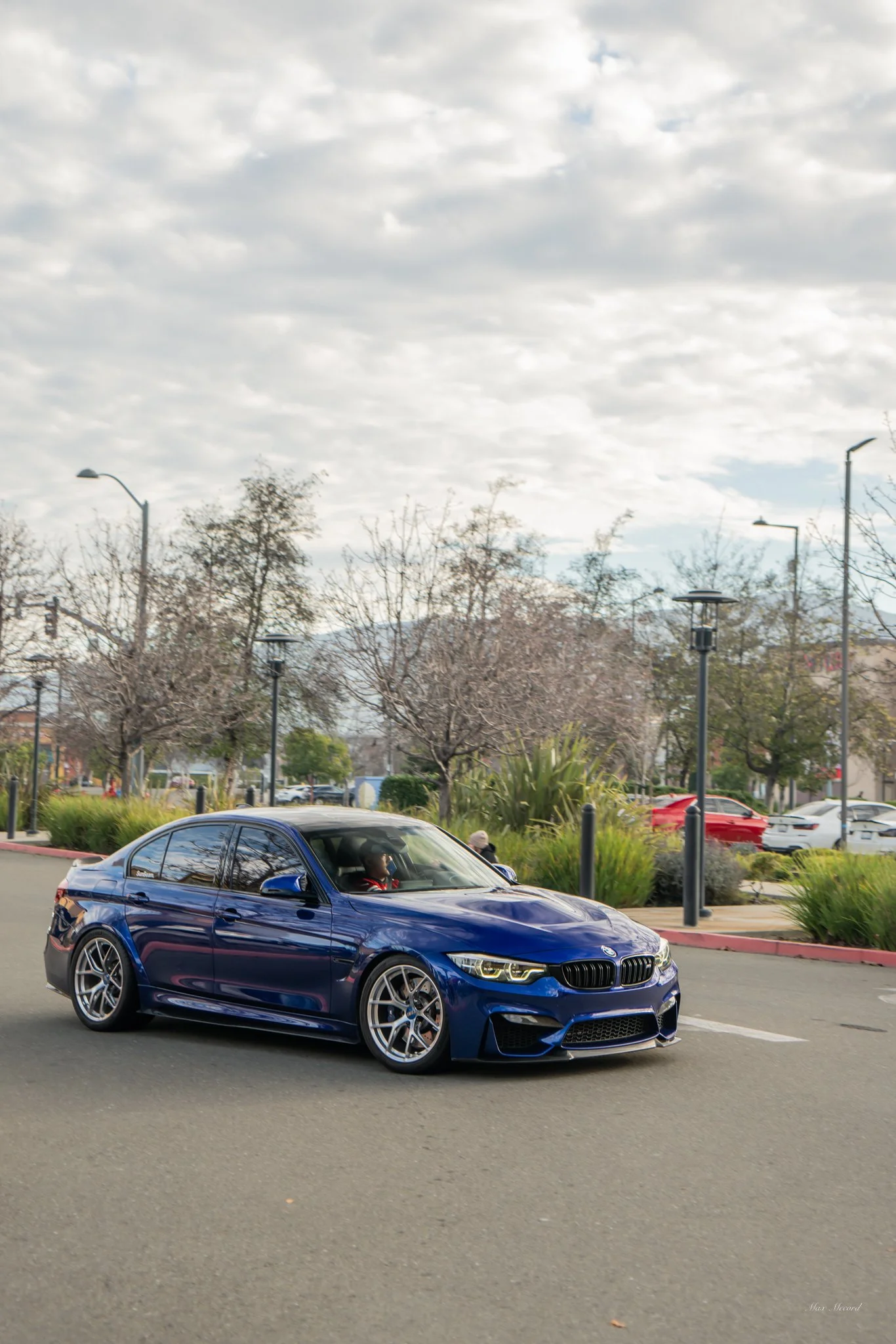 A blue BMW M3 sports car driving on a city street with trees, street lamps, and parked cars in the background.