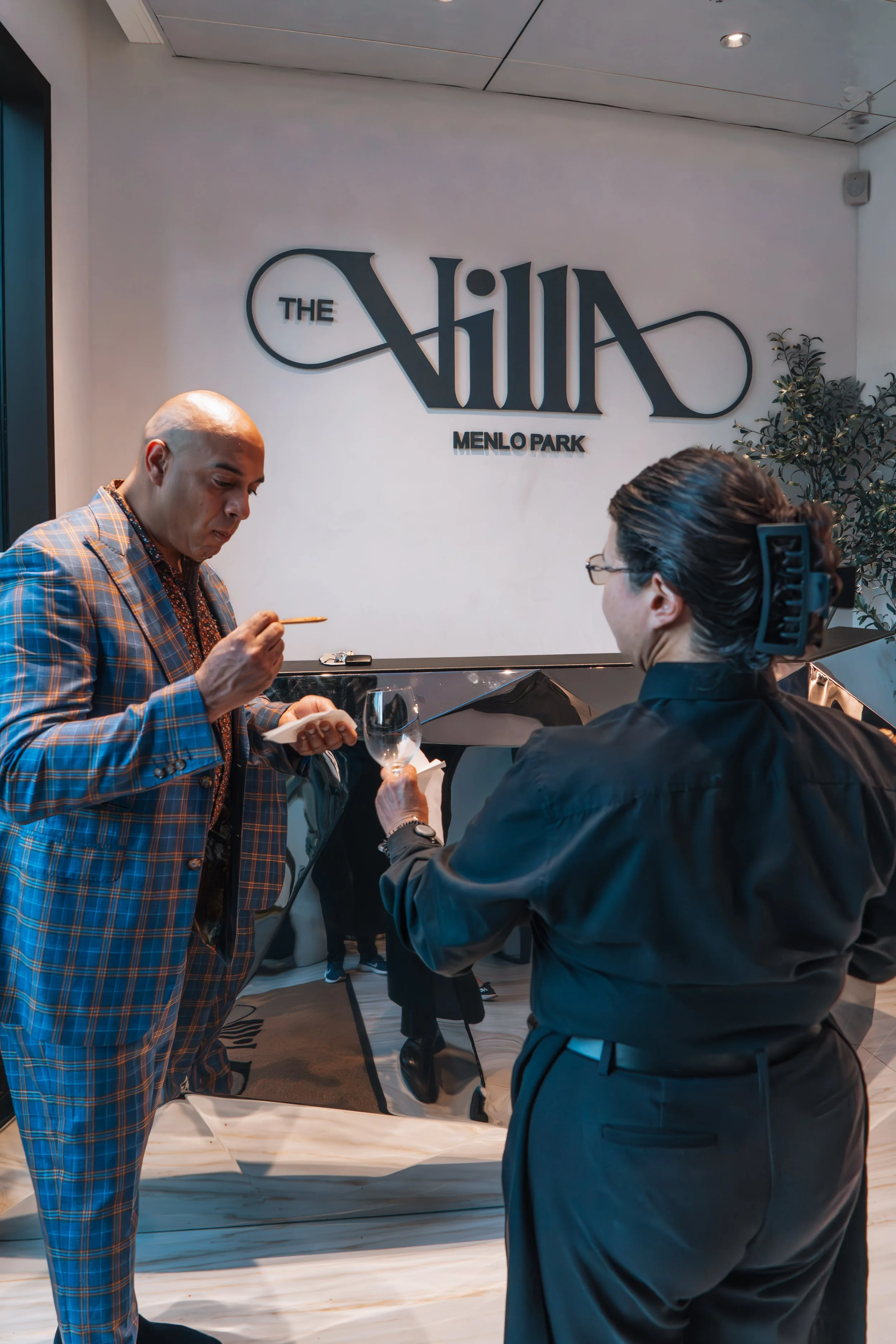 A man in a plaid suit uses a toothpick while talking to a woman in formal attire holding a wine glass, inside a modern upscale restaurant with the sign 'The Villa Menlo Park' on the wall.