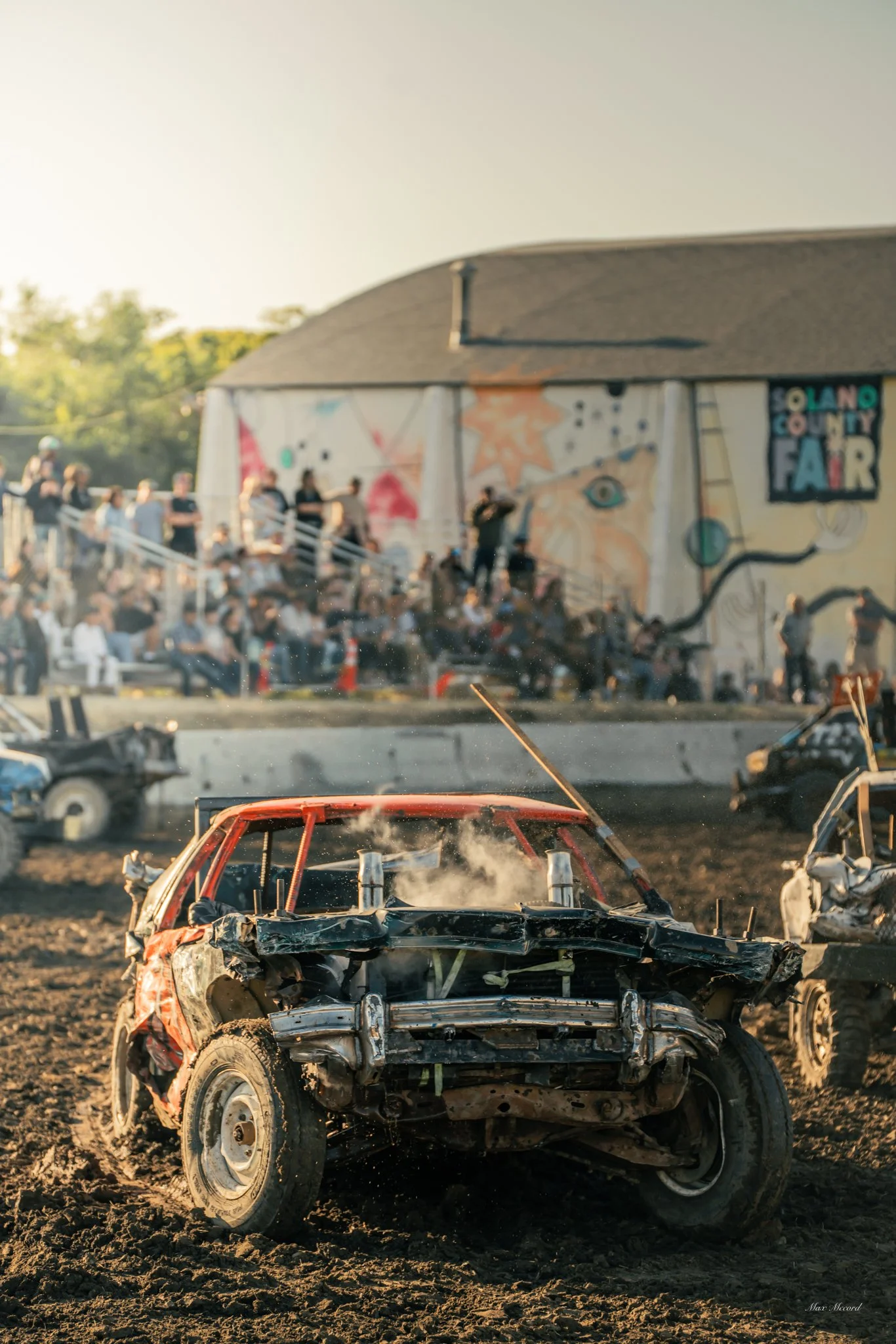 A damaged car on a dirt track at the Solano County Fair with a crowd of spectators and a mural in the background.