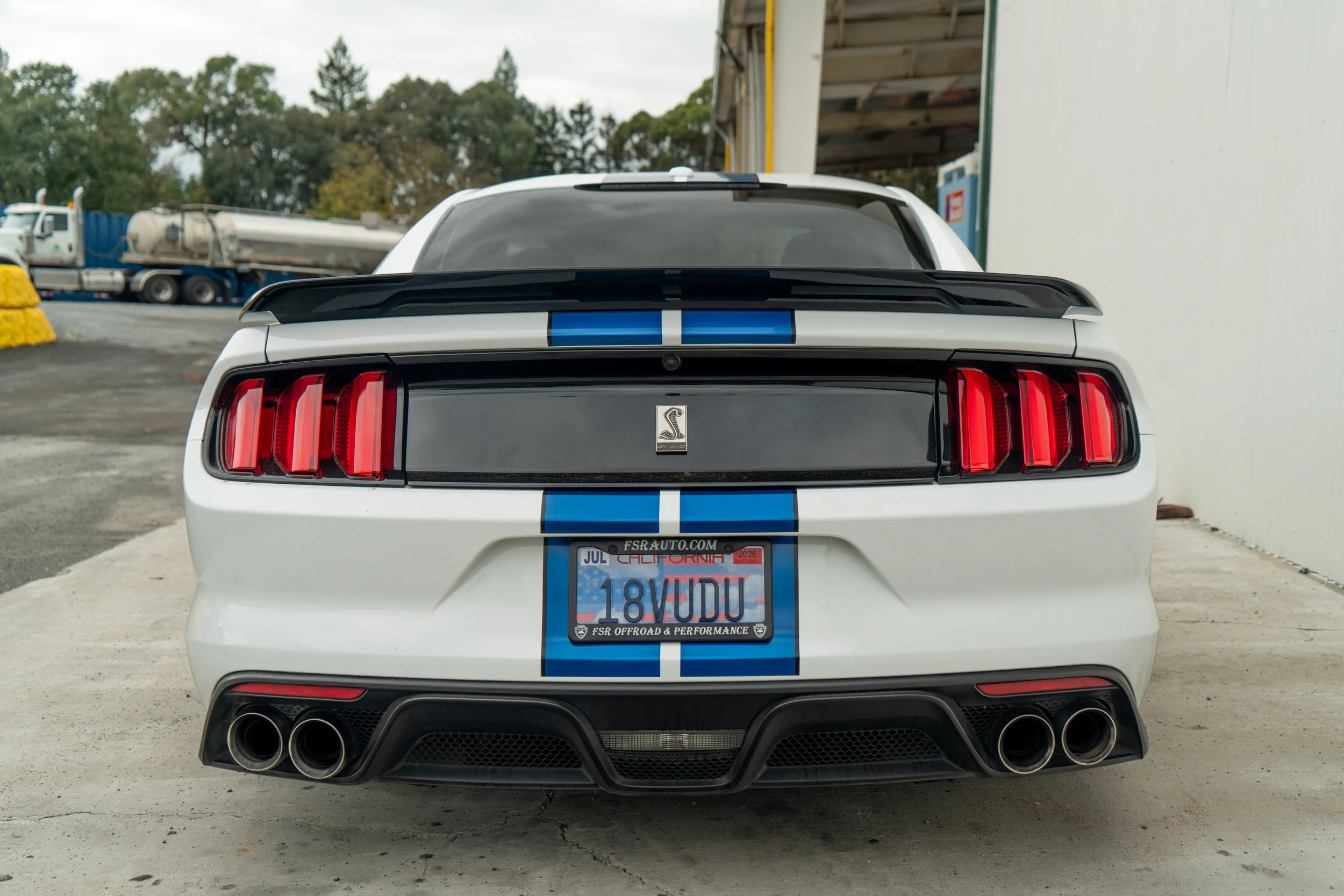 Rear view of a white Ford Mustang Shelby GT500 with blue racing stripes, black spoiler, quad exhaust pipes, and California license plate reading 18VUDU.