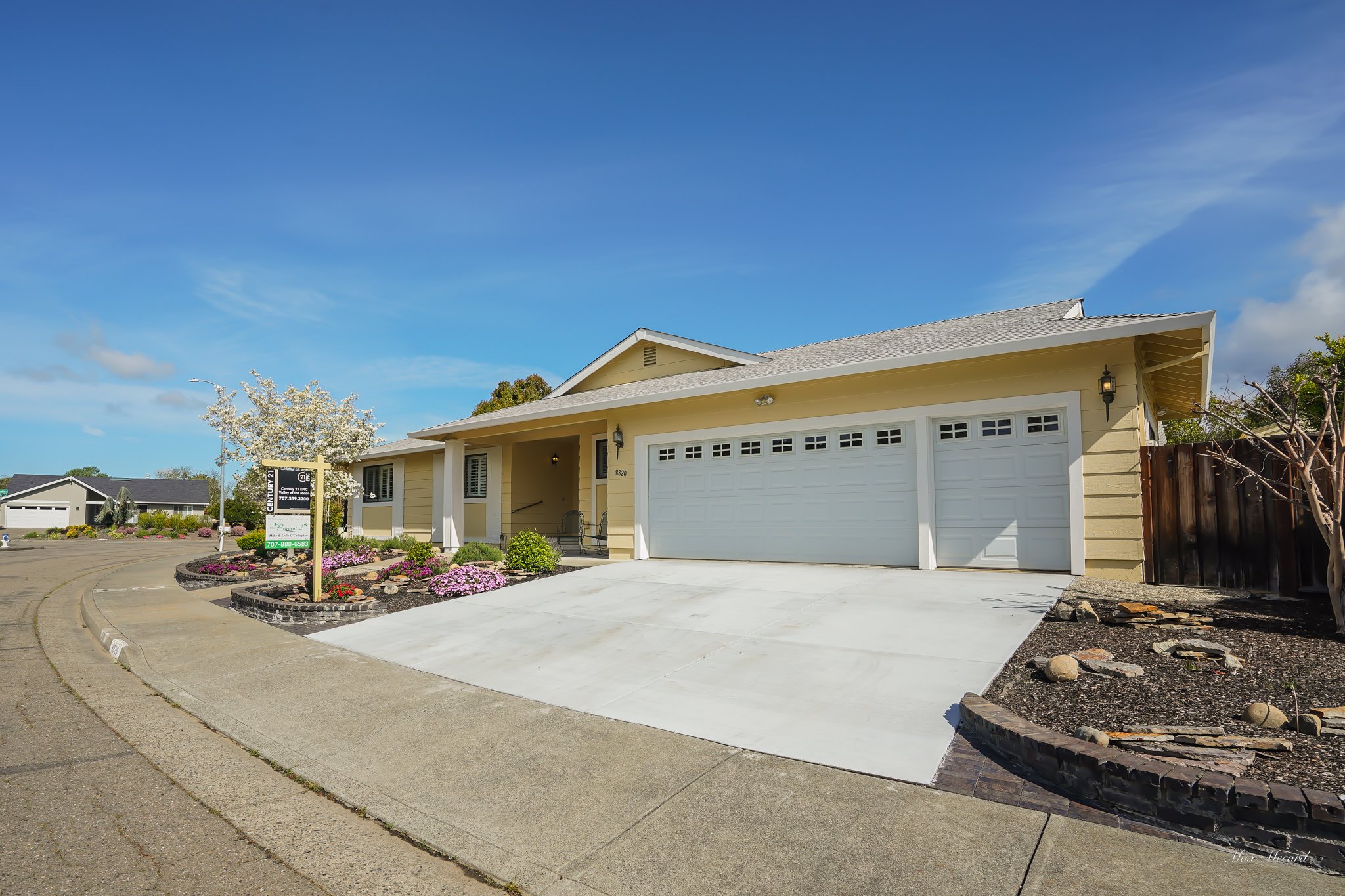 Single-story yellow house with white garage doors, a front porch, and landscaped front yard with pink and purple flowers, a white flowering tree, and a sidewalk.