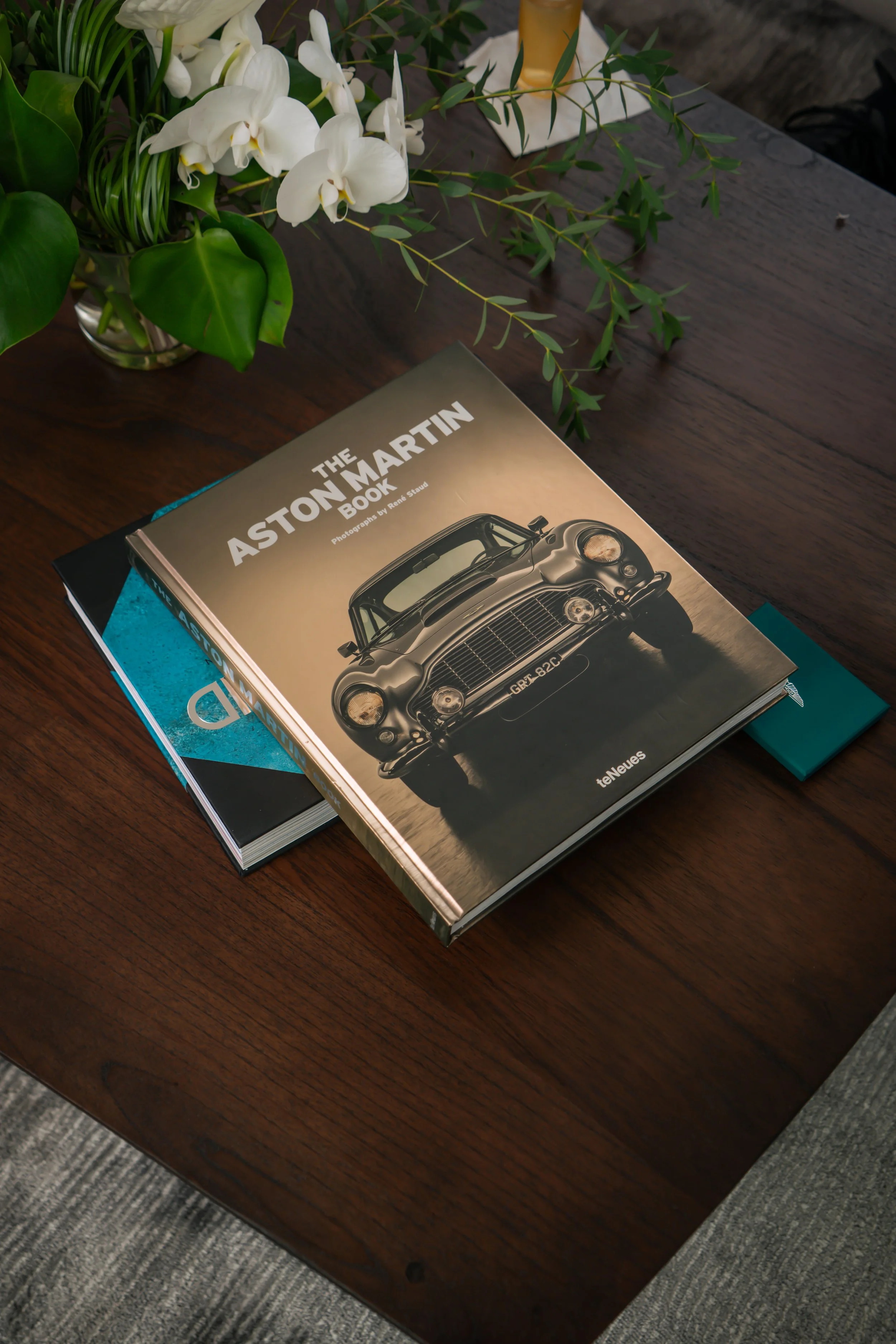 A book titled 'The Aston Martin Book' featuring a classic Aston Martin car on the cover, placed on a wooden table next to a vase of white orchids and green foliage.