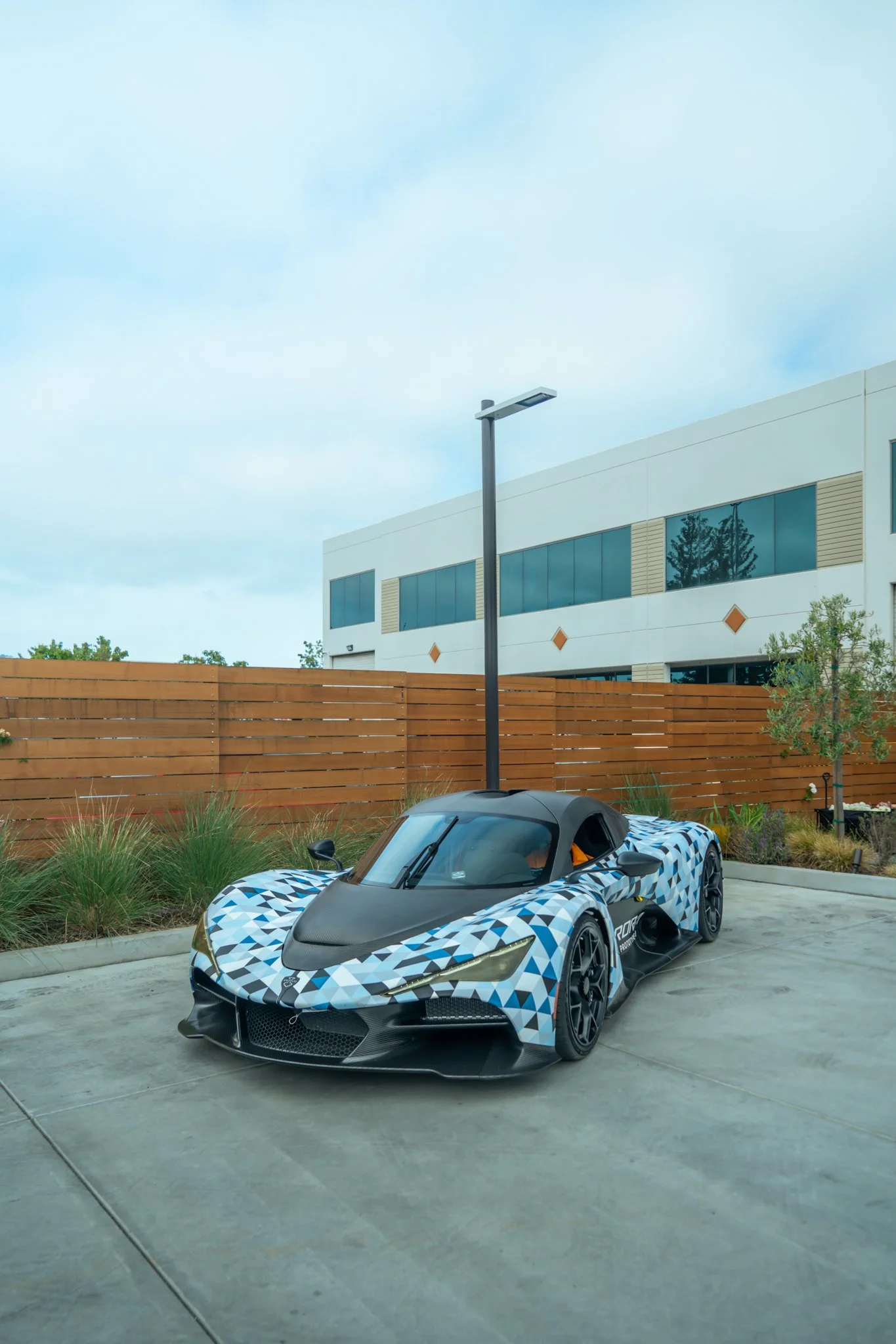 A race car with a geometric blue, black, and white pattern on a concrete surface in front of a wooden fence and modern building.