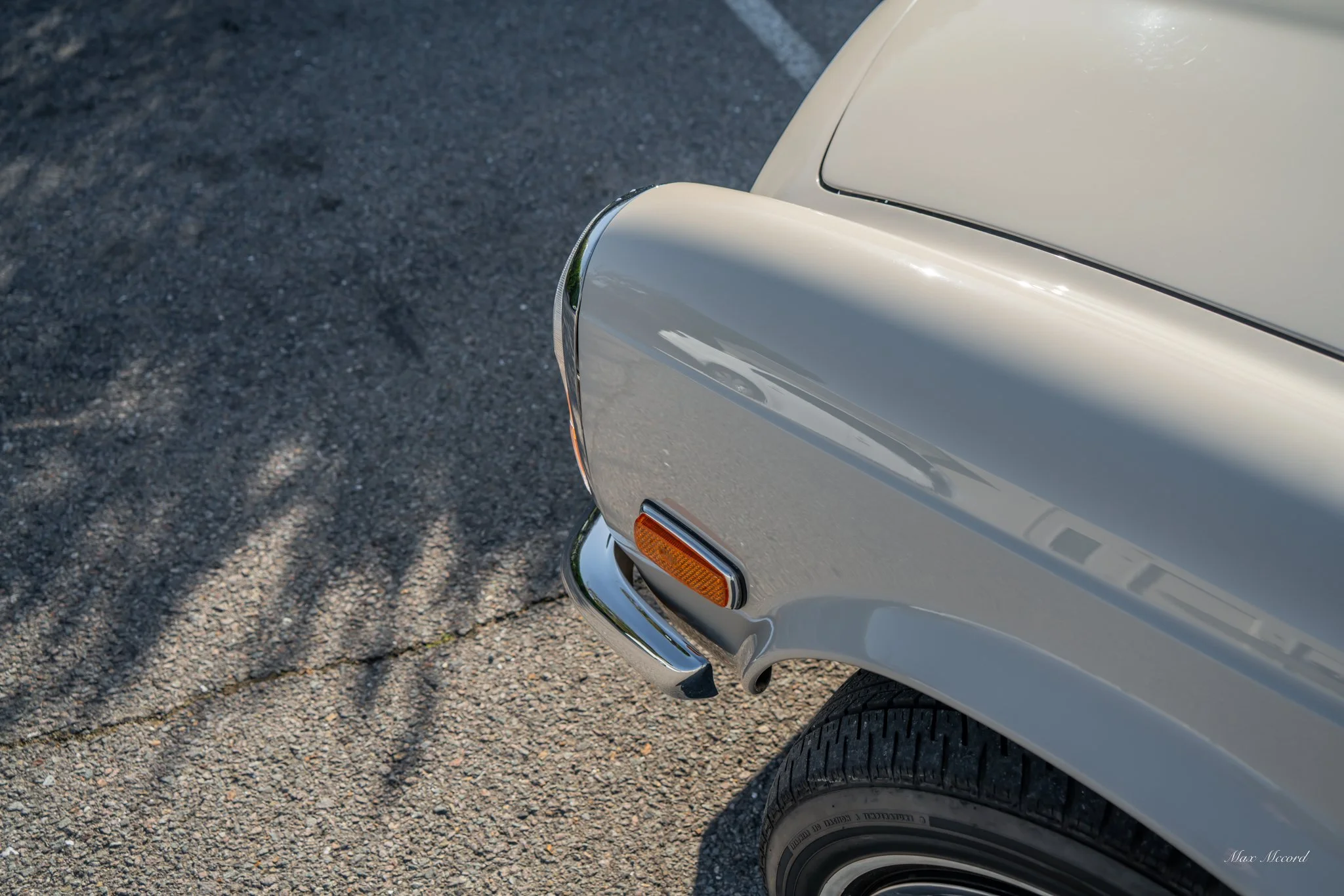 Close-up of the front right corner of a vintage silver car, showing part of the tire, a chrome bumper, and an orange turn signal light, parked on a gravel driveway.