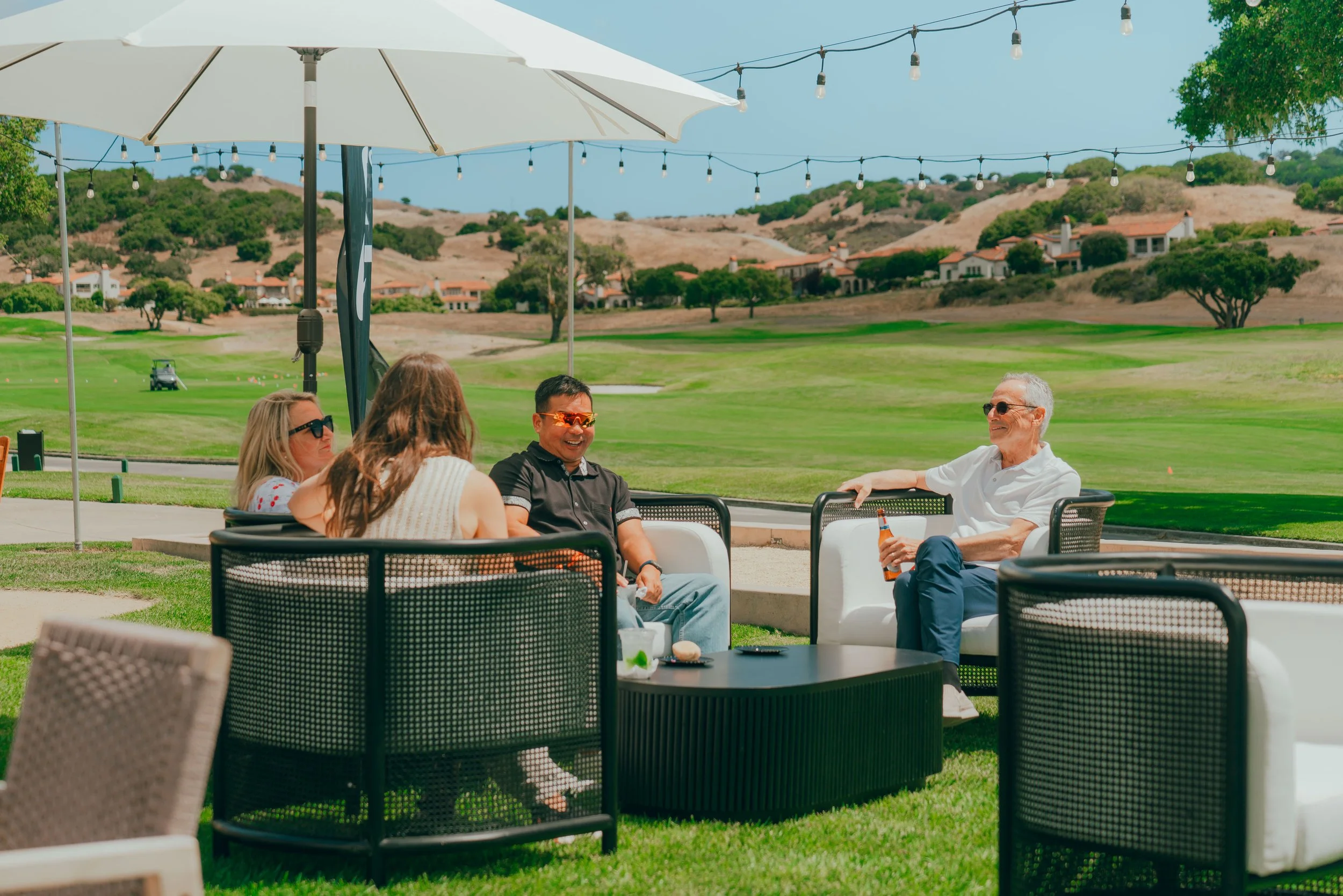 Four people sitting outdoors at a lounge area on a golf course, enjoying drinks and conversation on a sunny day with string lights and scenic hills in the background.